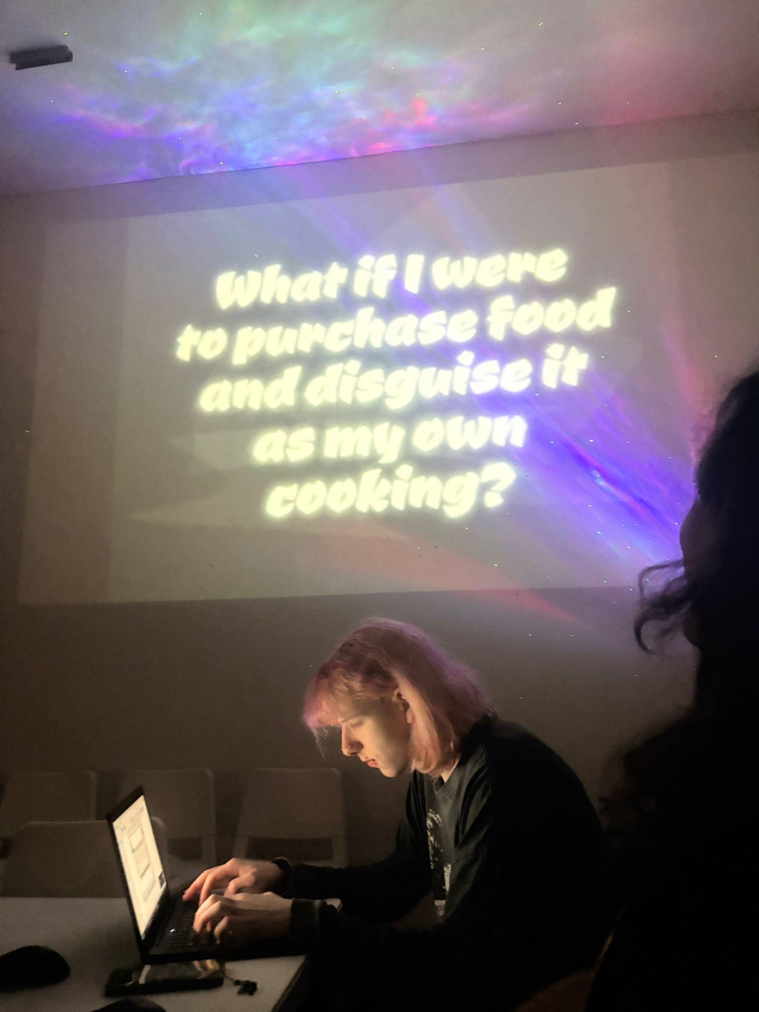 A person typing at a laptop in front of a screen with text 'what if I were to purchase food and disguise it as my own cooking'