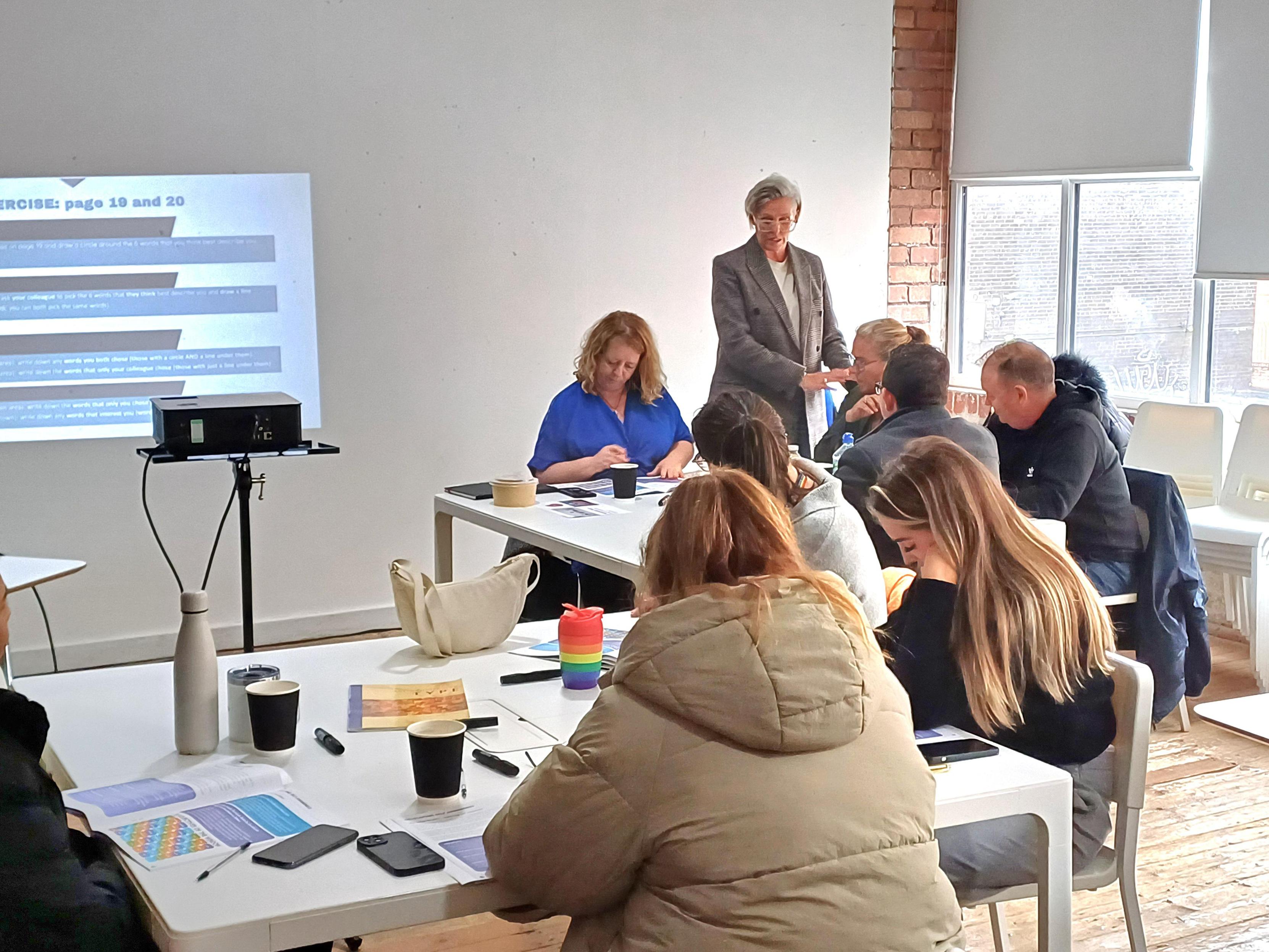 People sitting at tables with workbooks and drinks in a room with white furniture and blinds and a wooden floor, watching a presenter present slides