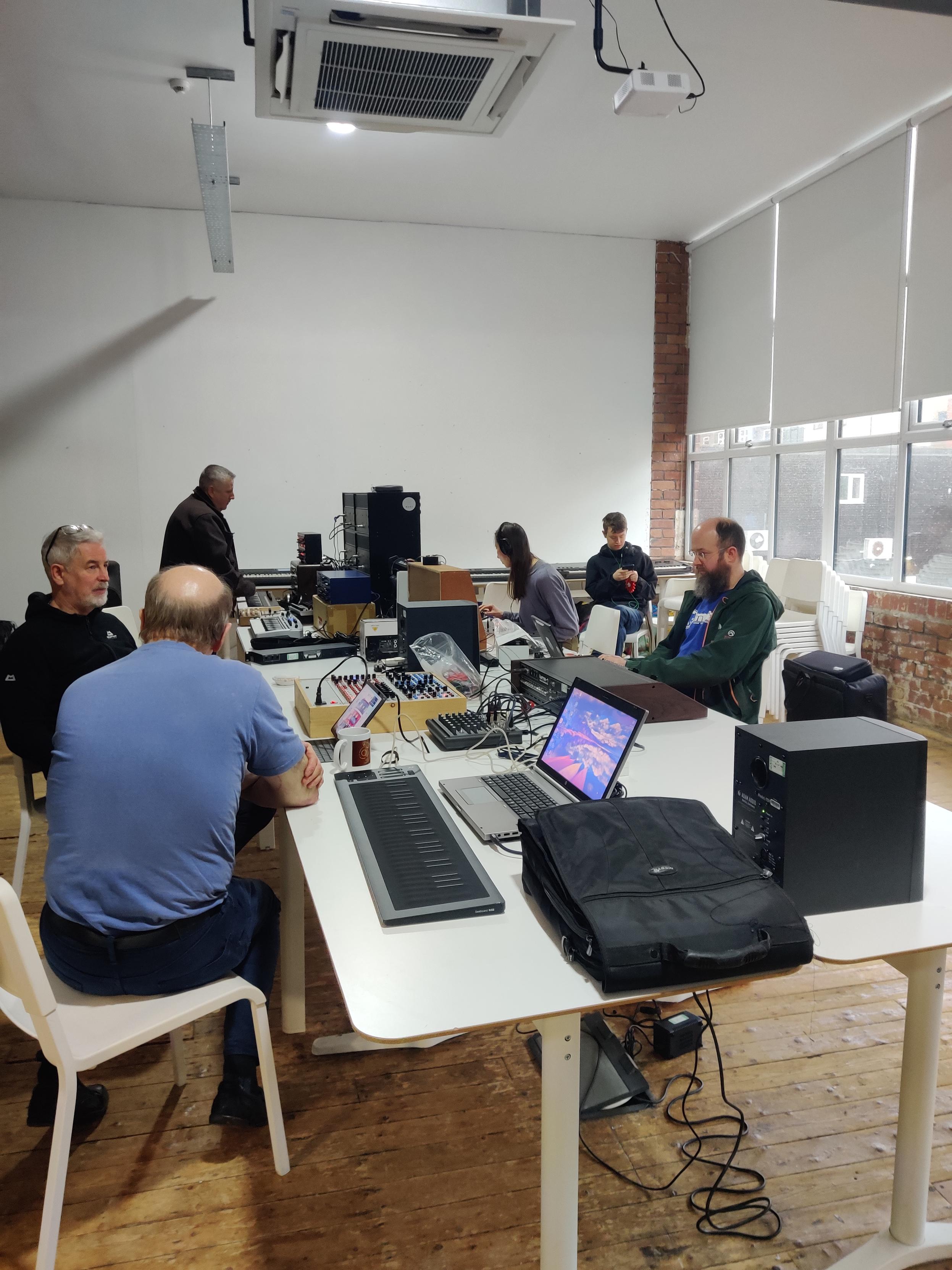 A group of people sat around a white table with synths and laptops