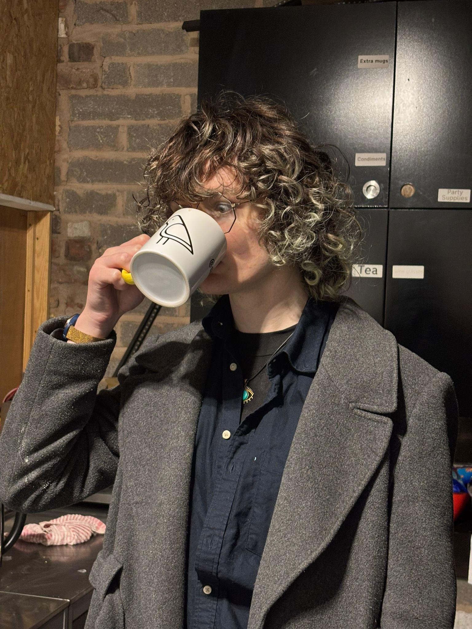 Poppy, standing in the DoES Liverpool kitchen, drinking from a mug with the Shelf Games logo