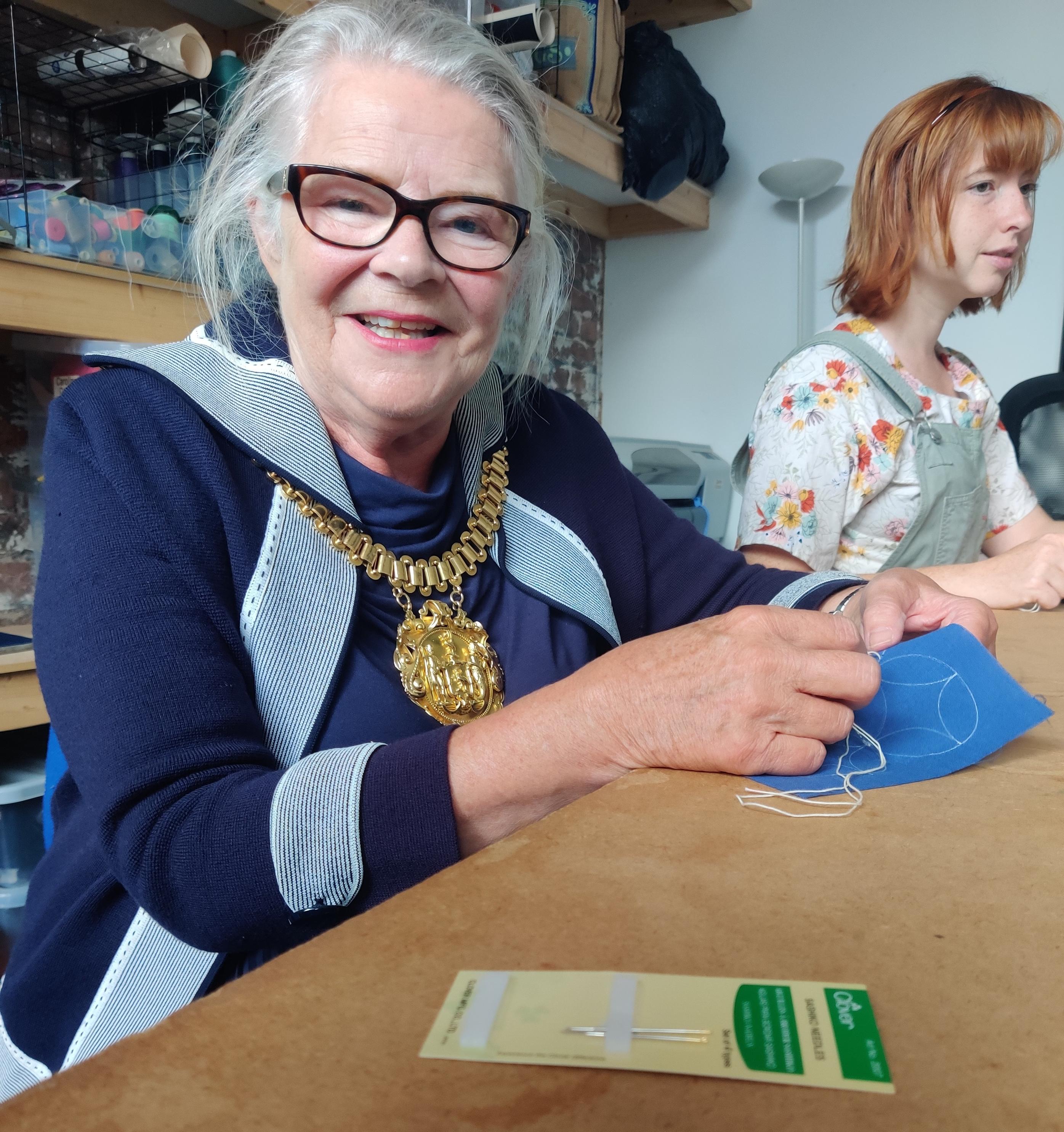 Lord Mayor Barbara Murray sitting by a table and sewing a sashiko patch, AJ in the background 