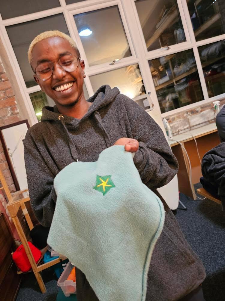 Dahab in front of a window in the DoES Liverpool Fibre Arts room, smiling, holding up a piece of light blue fabric with a star embroidery in yellow and green