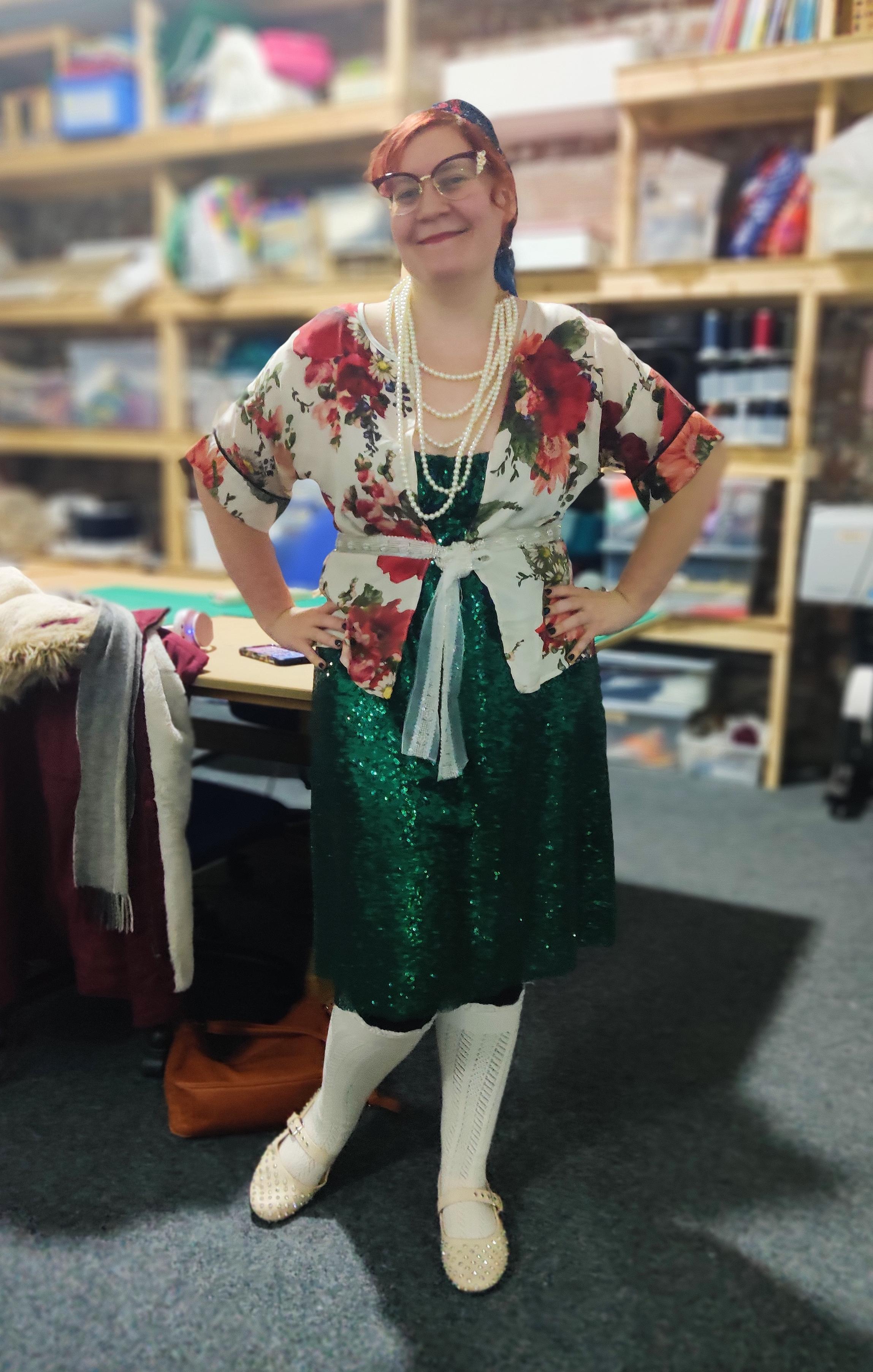 Jamie, standing in our Fibre Arts room, wearing pearls, a rose-patterned white jacket with white fringed tie at the waist, green sequin dress, white patterned ties and white sequin Mary Jane shoes