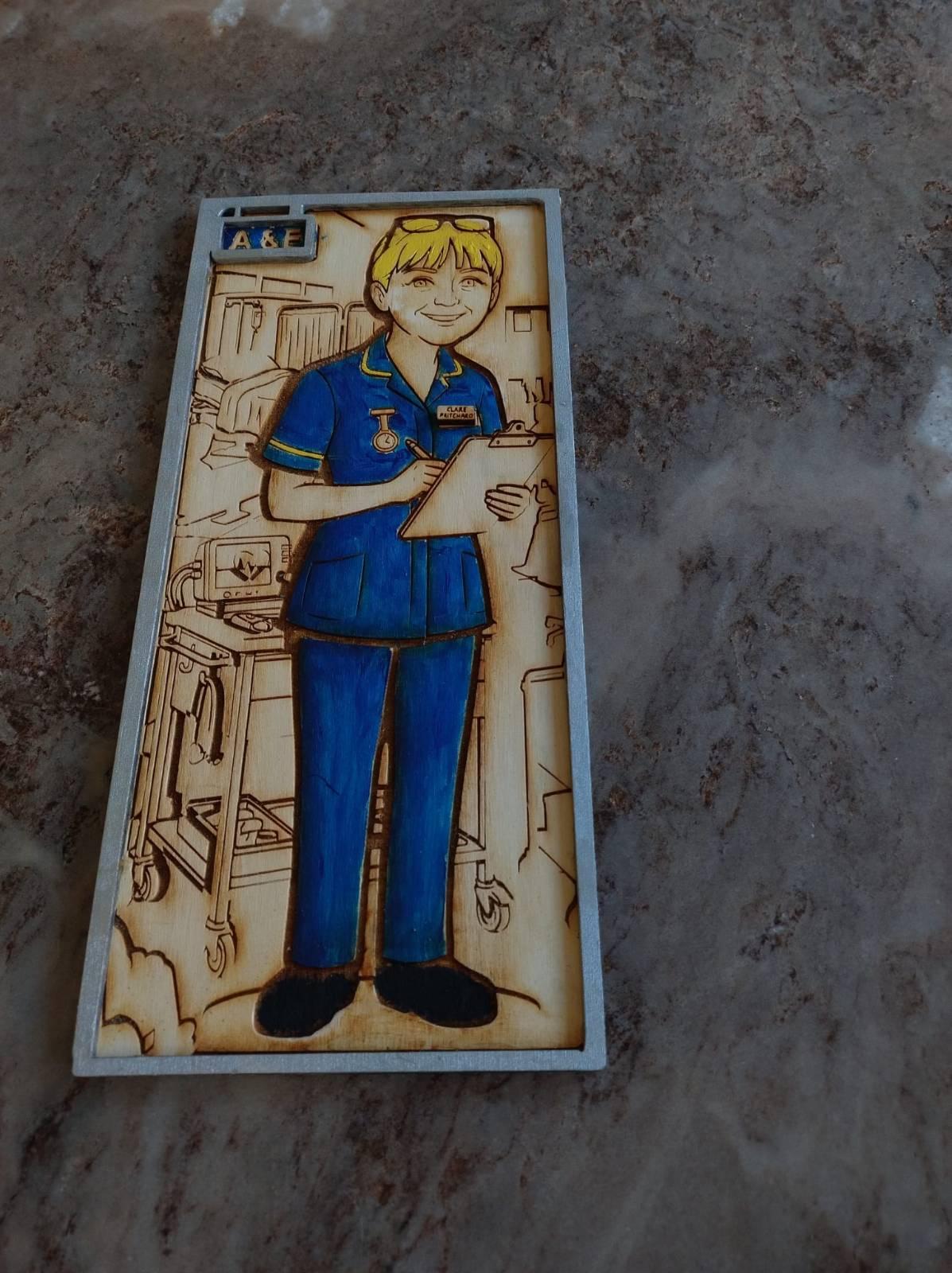 Photo of a laser cut, engraved and hand painted wooden plaque, showing a stylised picture of a nurse standing in front of a hospital bed.