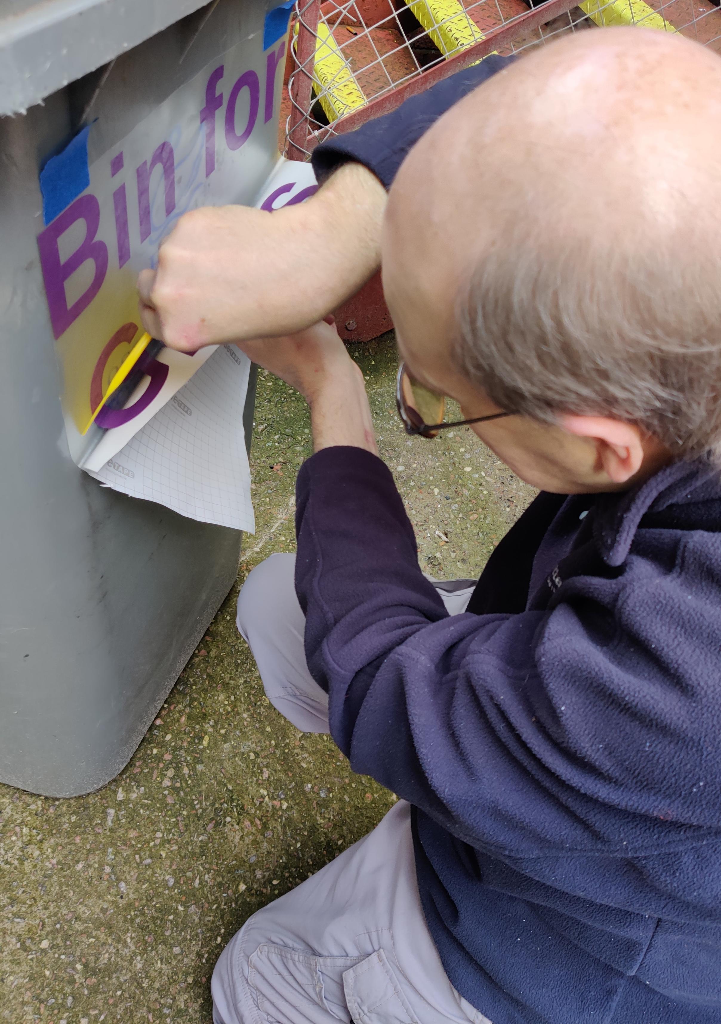 Mike G using a scraper to apply purple vinyl lettering to a grey wheelie bin