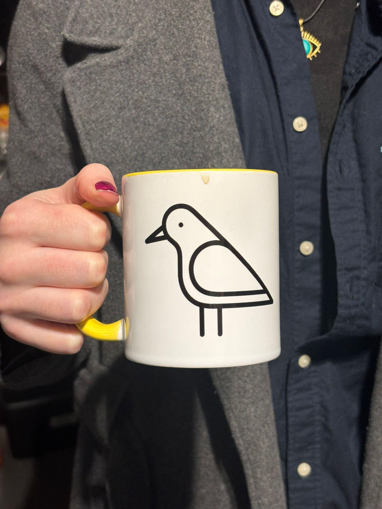 A close-up of the Shelf Games mug, featuring a black stylised outline bird, in black on a white background. The mug has a yellow handle and inside