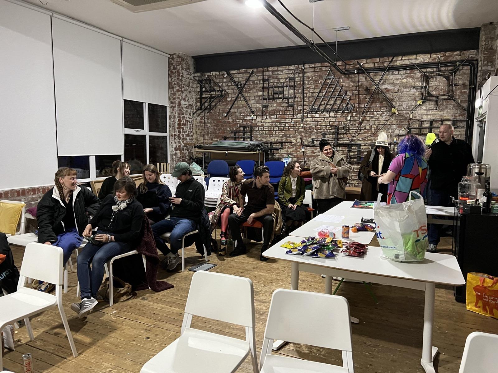 The DoES Liverpool Event Space, with white and brick walls and a wooden floor. People are sitting on rows on white chairs, and there are some white tables too 