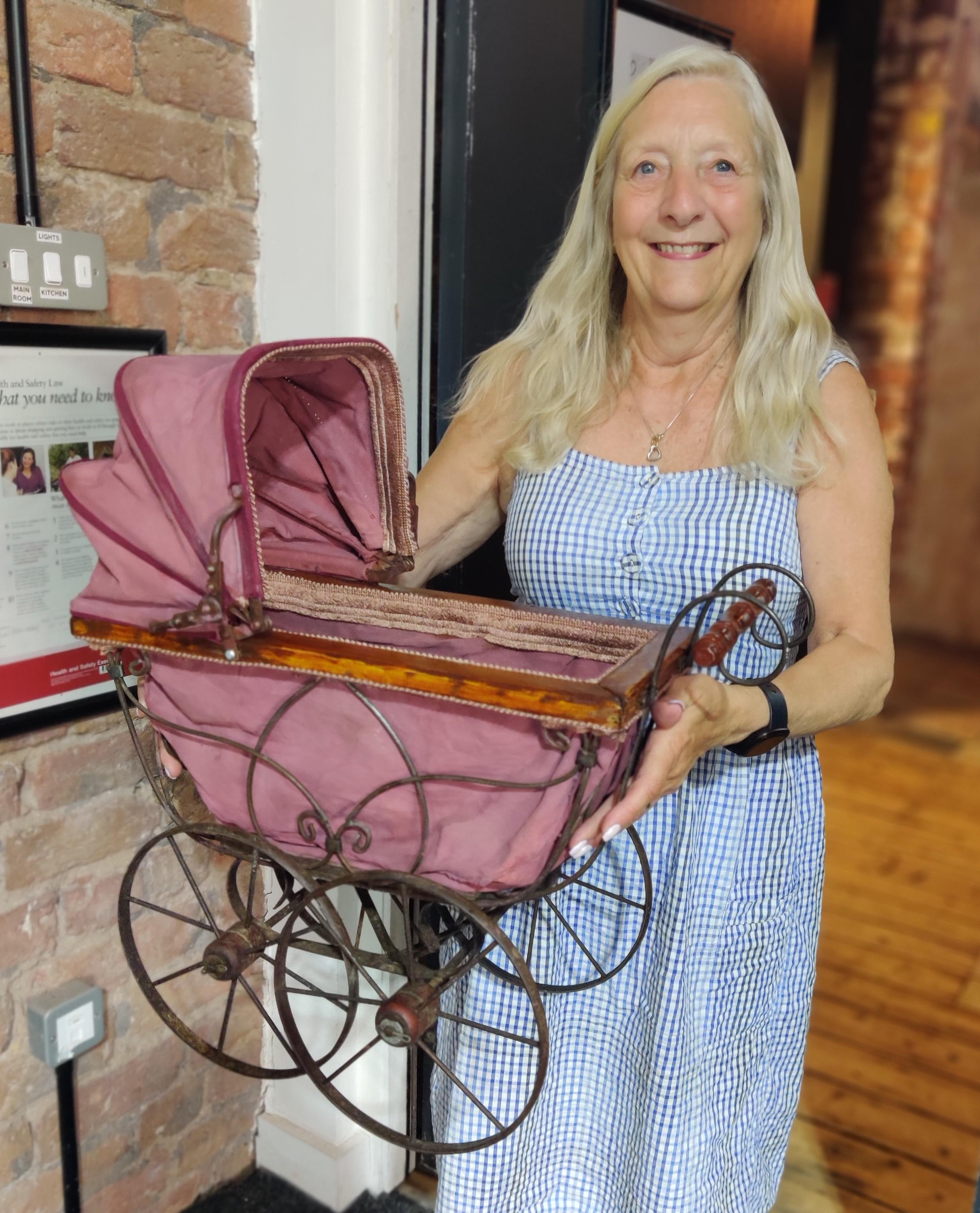 A light haired woman wearing a sundress and holding up an sntique-looking toy pram.
