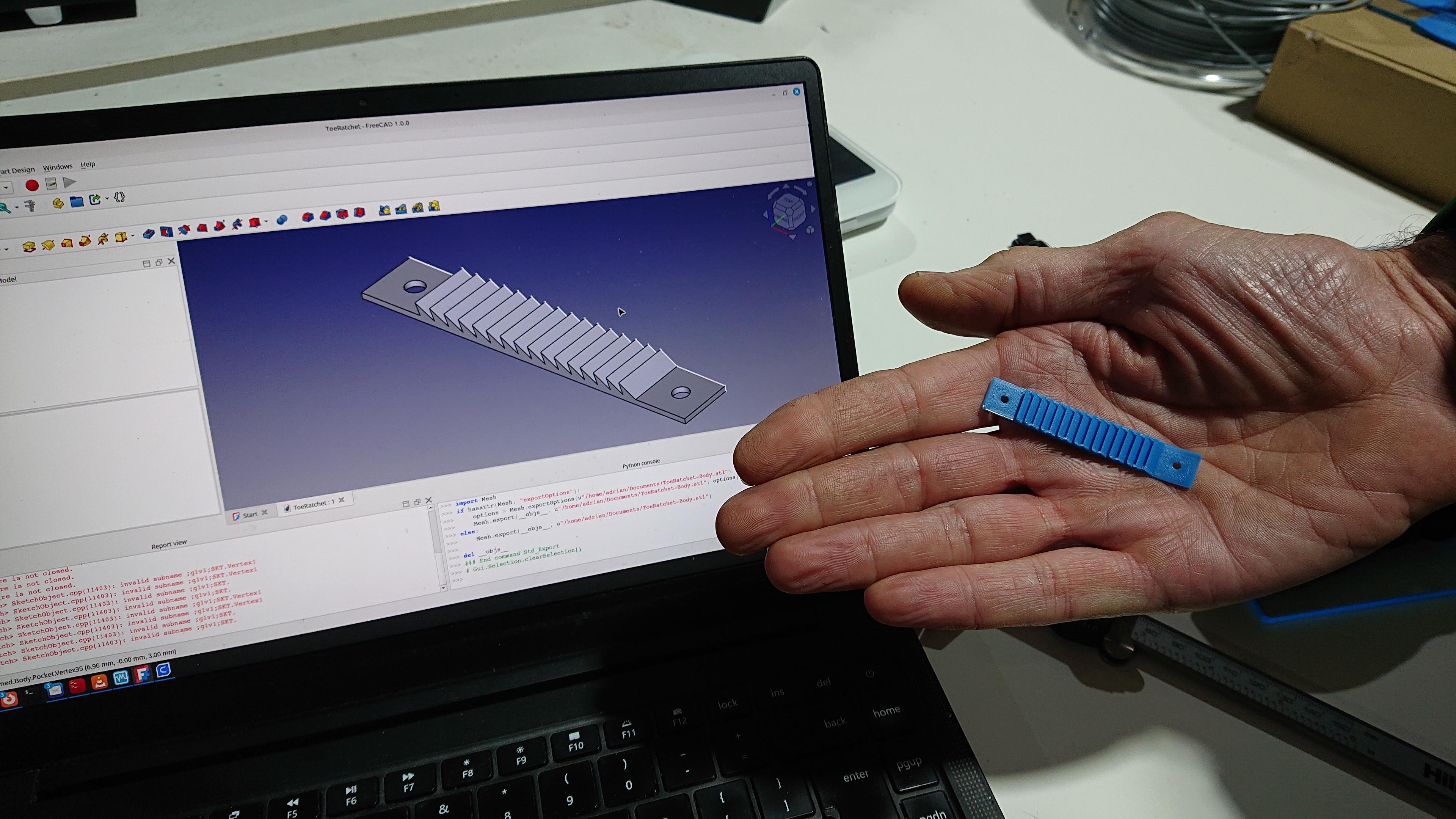 A person's hand is shown holding a small blue 3D print of a toothed ratchet with screwholes at either end. Behind their hand is a laptop showing a 3D render of the same model from where it was designed in the FreeCAD software