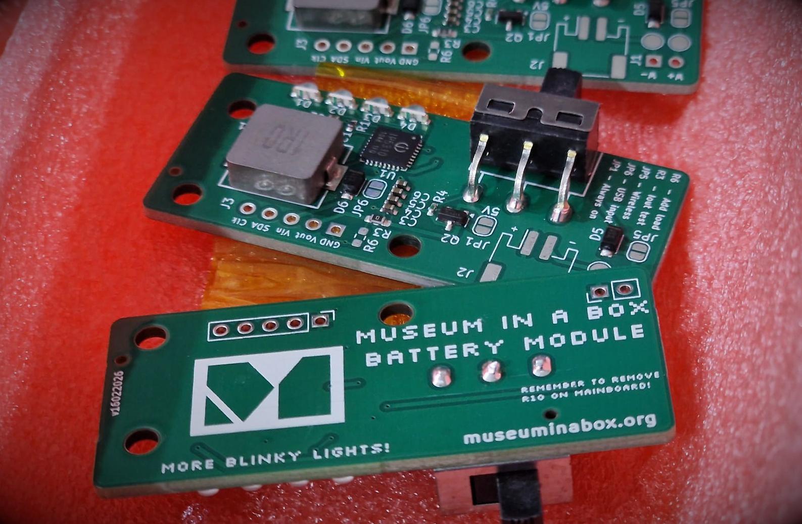 Three, of five, green PCB's that allow for battery power. The foreground shows the front with a Museum in a Box logo and the text 'Museum in a box battery module' and 'More blinky lights' The other boards show the reverse with a chunky on off switch, some leds and a battery power management ic.