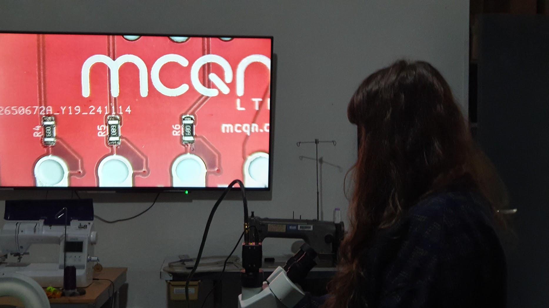 Mally is seen standing on the right of the photo, she is using a microscope on the table in front of her. The image from the microscope is projected on a large TV in the background. The PCB is seen with the letters MCQN and soldered components.