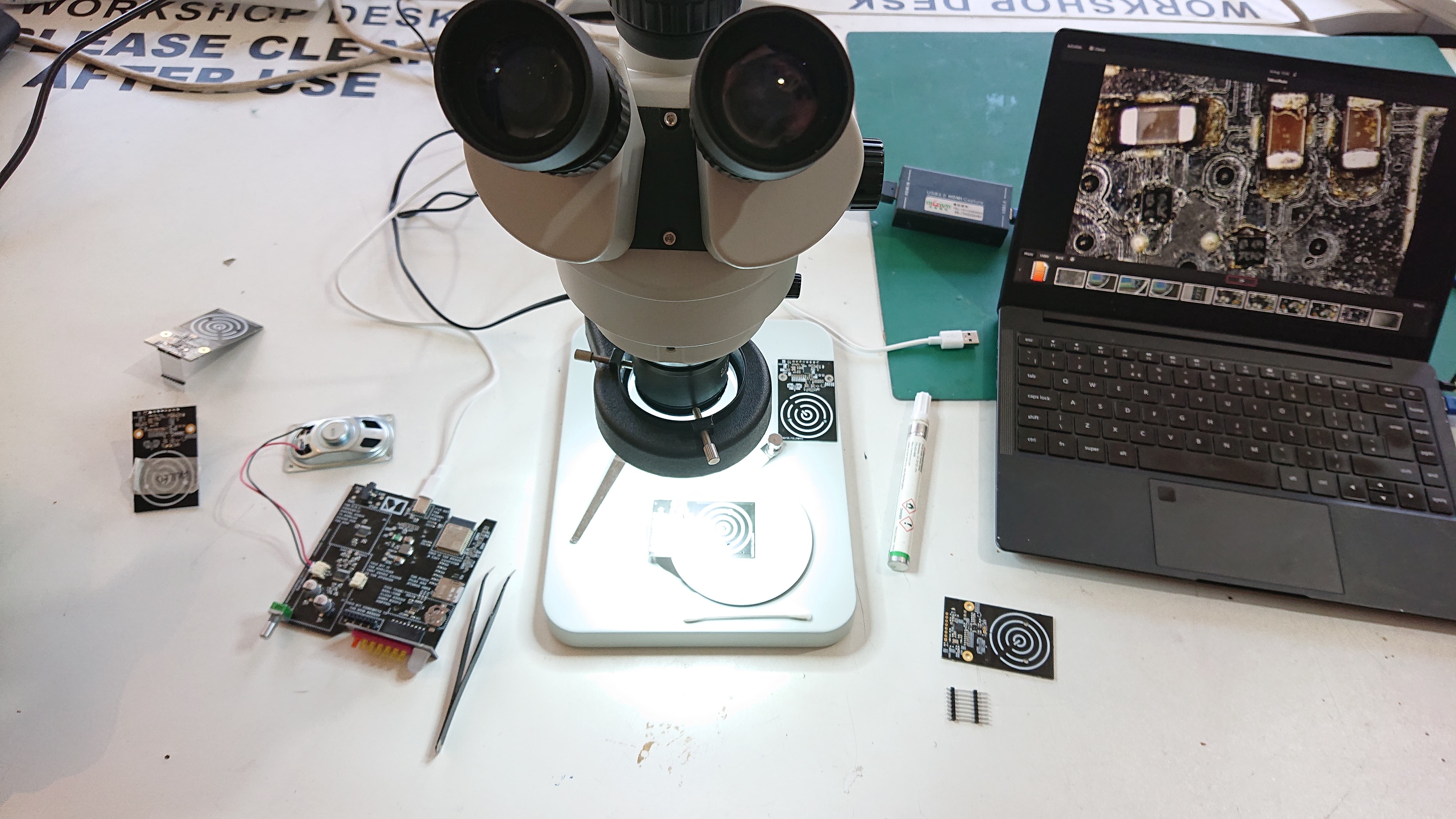 Looking down on a white desk that has a microscope on it and a laptop to the right.

The laptop screen is showing a picture of the view from the microscope and arrayed around the scope are some circuit boards, forceps, solder flux pen and cotton bud