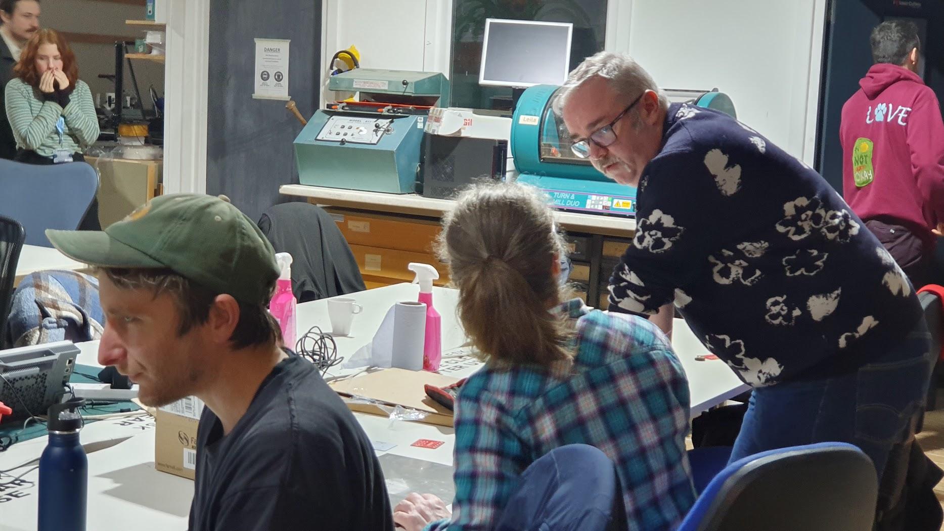 Adrian standing but leans on the table talking to Karen before the start of the PCB soldering workshop. In the foreground looking left is a man wearing a baseball hat, in the background behind Adrian is a man facing away in a red hoody with the word love on the right shoulder.