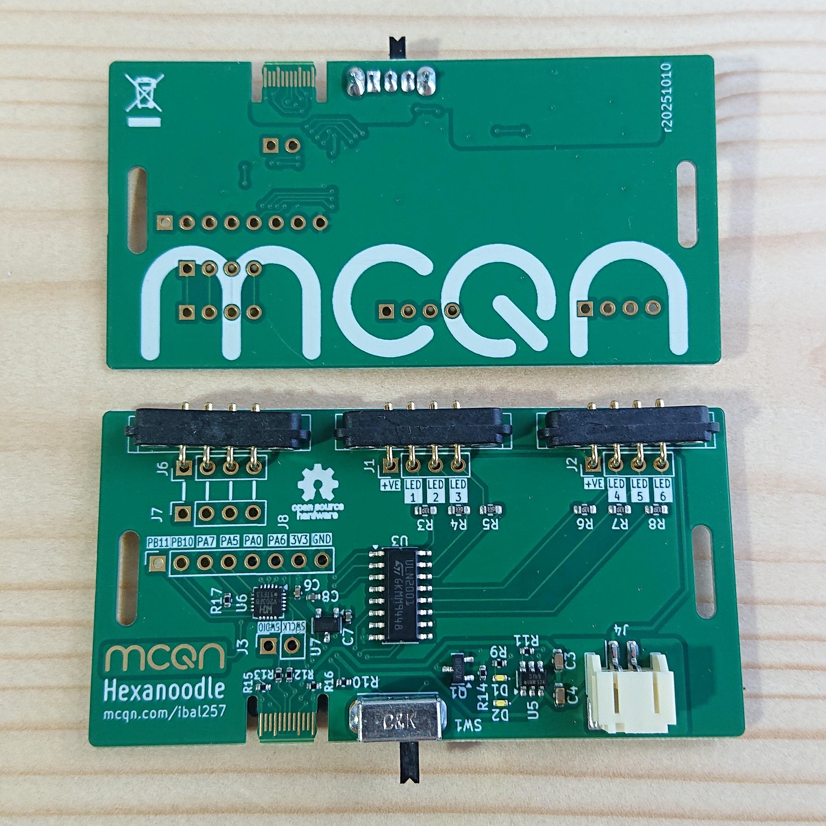 Two rectangular printed circuit boards sat on a wooden table. There's two so you can see both sides of the board: the underside is emblazoned with the MCQN logo; the top-side is where the action happens. It has a few chips on it, a slide on/off switch, USB-C connector and rechargeable battery connector running along one side, and three 4-pin magnetic pogo-pin connectors running along the other side. In the bottom left corner of the top-side is a gold MCQN logo (which will, hopefully, work as a capacitive touch pad) above the text Hexanoodle and mcqn.com/ibal257