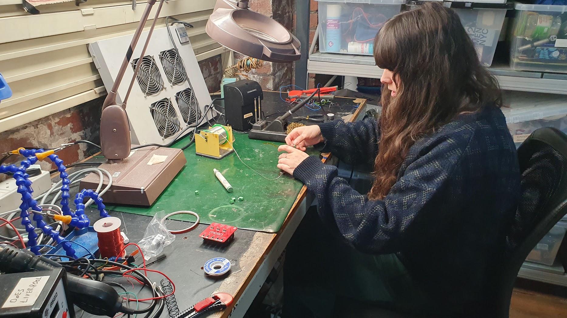 A young women Mally sits at a Soldering table spot welding items. She has long hair over her draped over her shoulders obscuring her face. Her hands are outstretched holding a PCB.