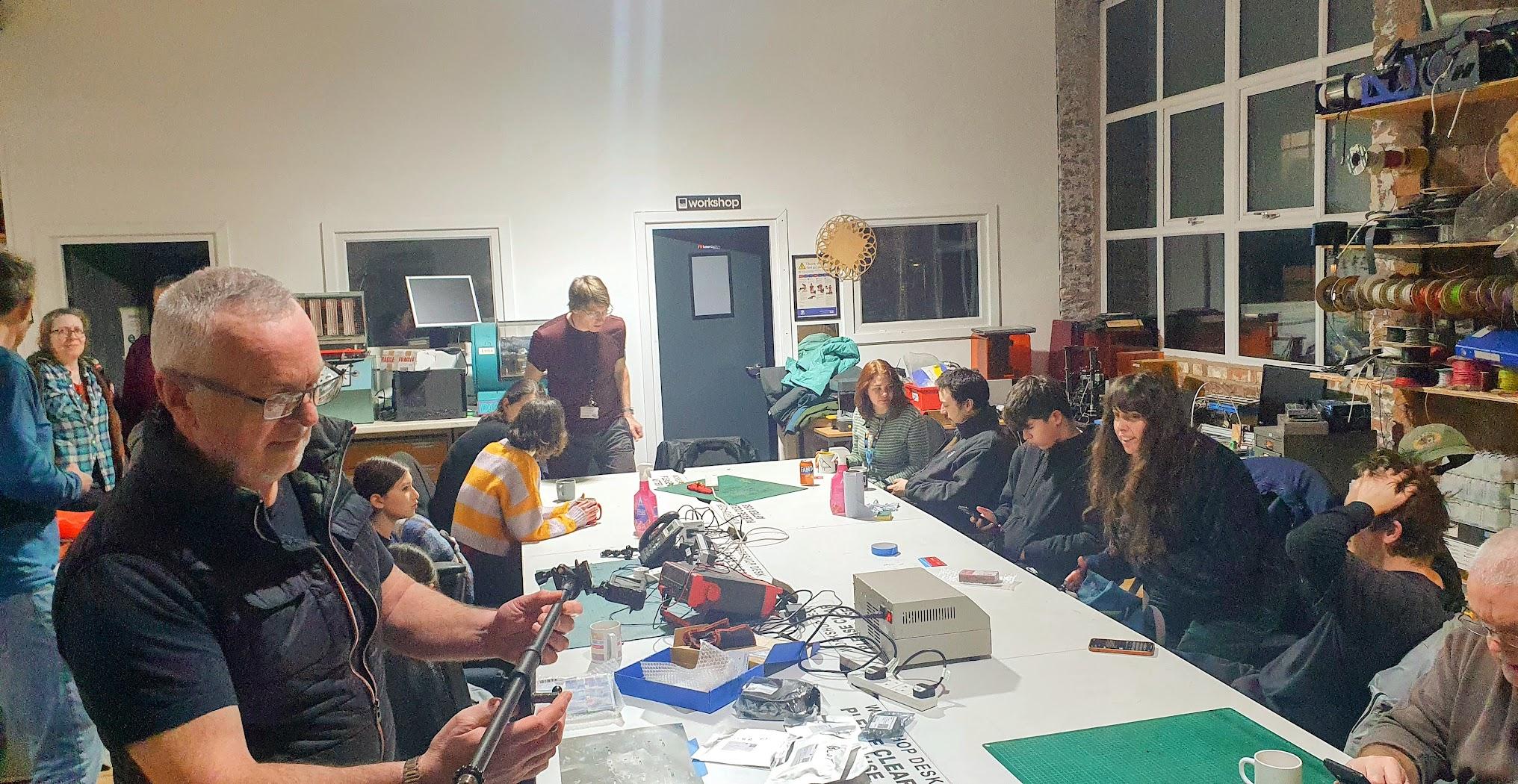 A man stand in the foreground manipulating a microphone stand in the background is a large white table - the table contains various electronic equipment. Around the table sit 11 people awaiting the soldering workshop to begin.