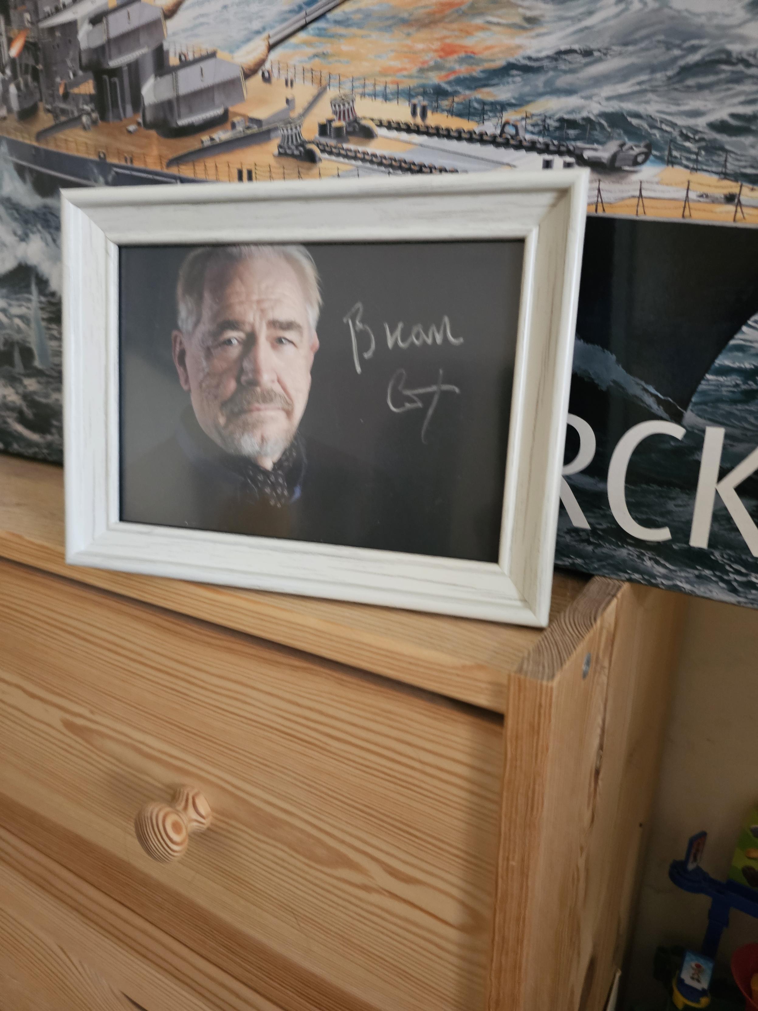 A signed photograph of Brian Cox, the actor, in a white frame atop a pine chest of drawers.