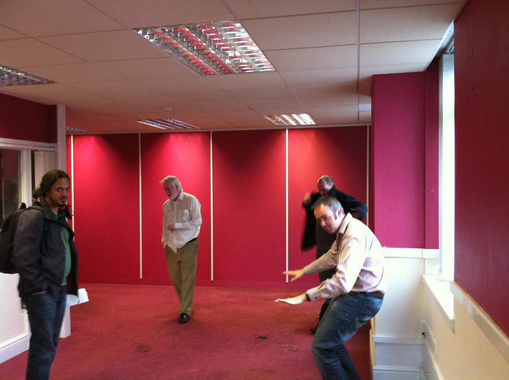 Four people in an empty office room with red walls and carpet. One of them, who looks a bit like me but can't be as he doesn't have a moustache..., looks like he's playing imaginary record decks while a couple of others look on in bemusement.