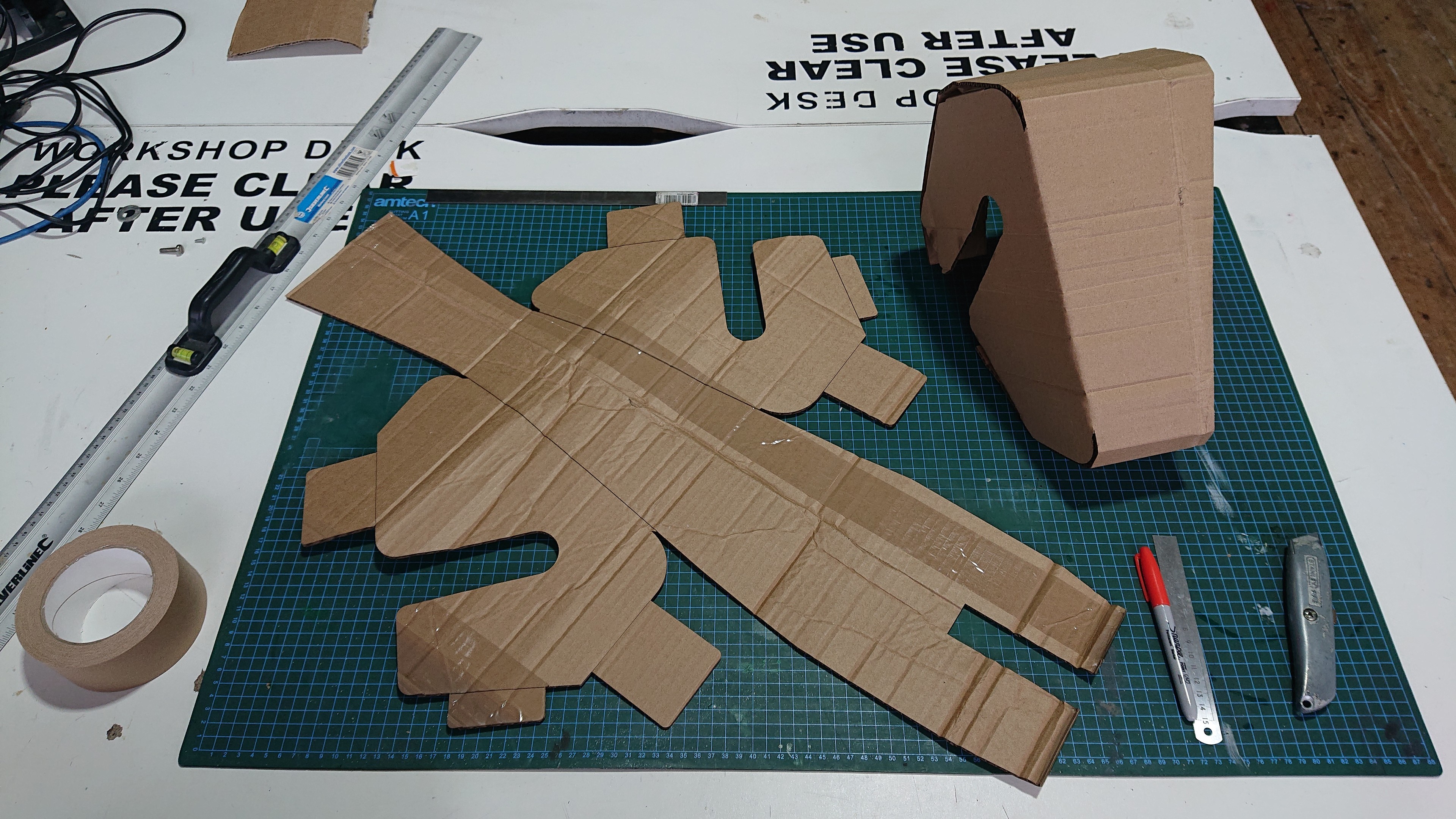 A workbench with a cutting mat, some tape, a sharpie, a Stanley knife and a metal ruler. On the cutting mat is a flat sheet of brown corrugated cardbaord cut out in the shape of the unwrapped mesh from the CAD design. Next to that is a version which has been folded up to make the triangular wedge shape.