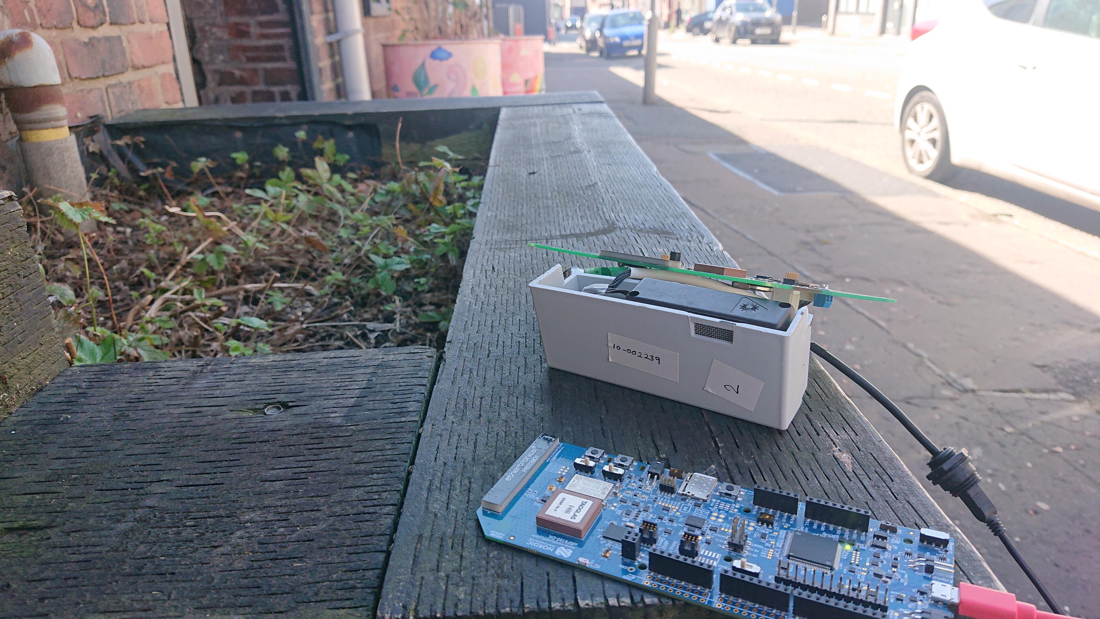 A couple of circuit boards are sat on the side walls of a planter on a city street. The remnants of the alpine strawberry plants are just visible in the planter