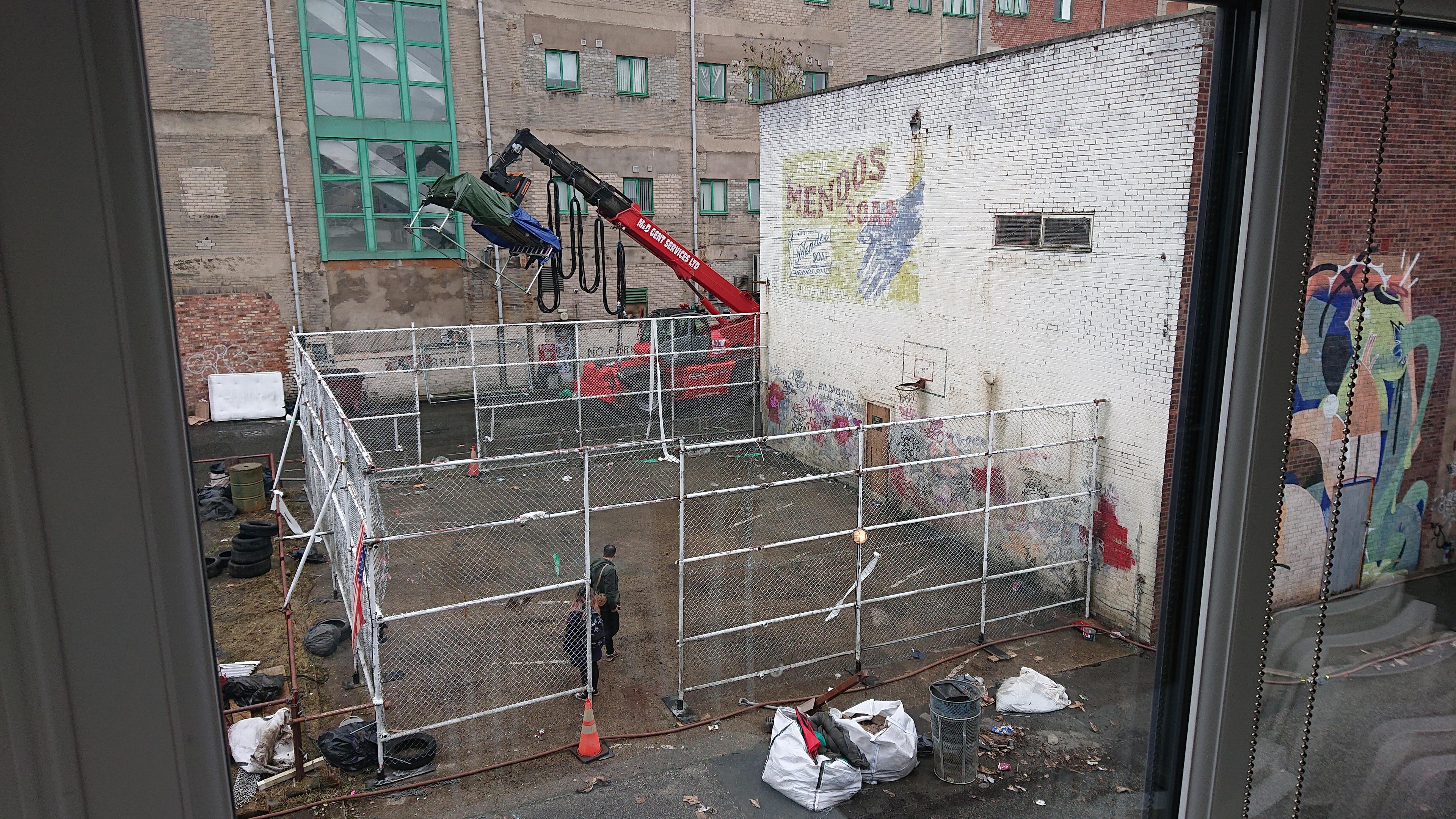 A half-basketball court with chain-link fencing round three sides; the fourth side is the wall of a brick warehouse. There's a faded sign for Mendos soap painted on the side of the building, and graffiti along the bottom of the wall; and bits of stained paintwork dotted here and there. All of that has been carefully painted by the set artists, and the whole fence has been constructed and painted over the last day or two. There's a firefighting hose running along the edge of the court, which is actually for creating the clouds of steam to make the area look like New York (well, Gotham City) and in the background is a large red teleloader which has a seriously large lighting rig on it.