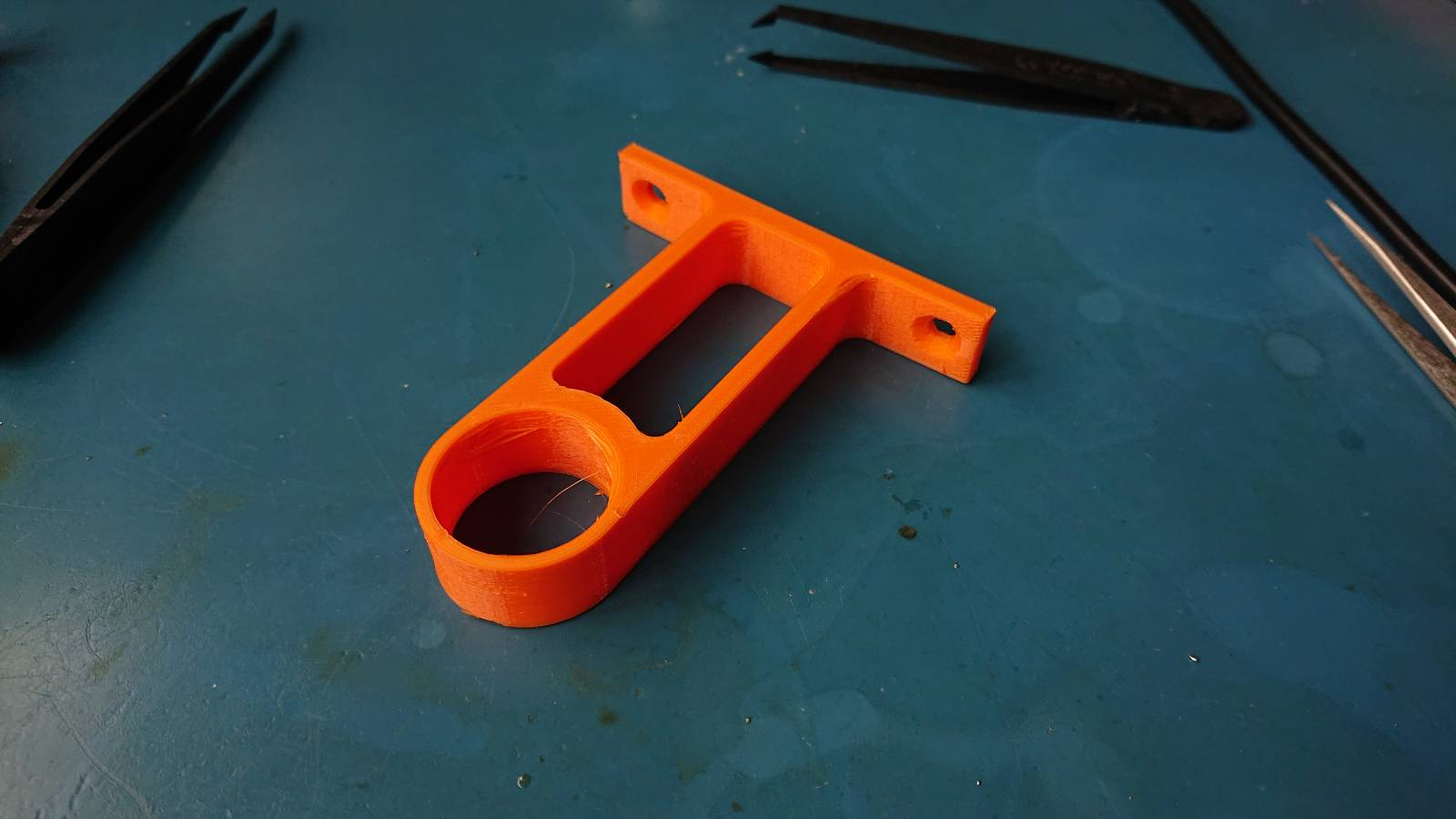One of the clips, 3D printed in orange PETG plastic, sits on a mat on a table next to a couple of pairs of forceps (they weren't involved in any of this, just happened to be lying around in the 