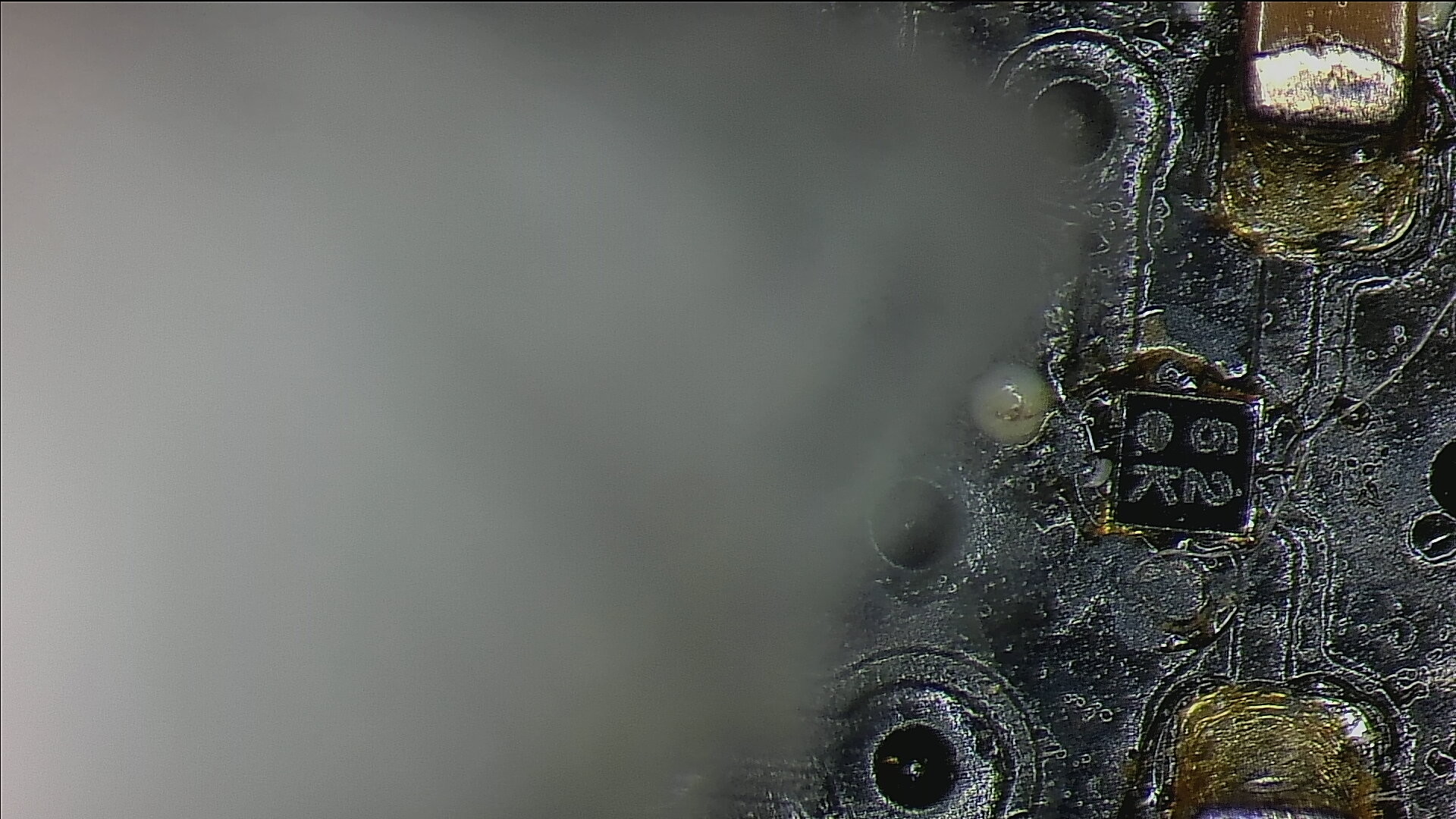 View of part of a circuit board through a microscope.  The main part visible on the board is a small black square with a couple of traces leading to it.

What looks like a large white cloud is obscuring most of the view.