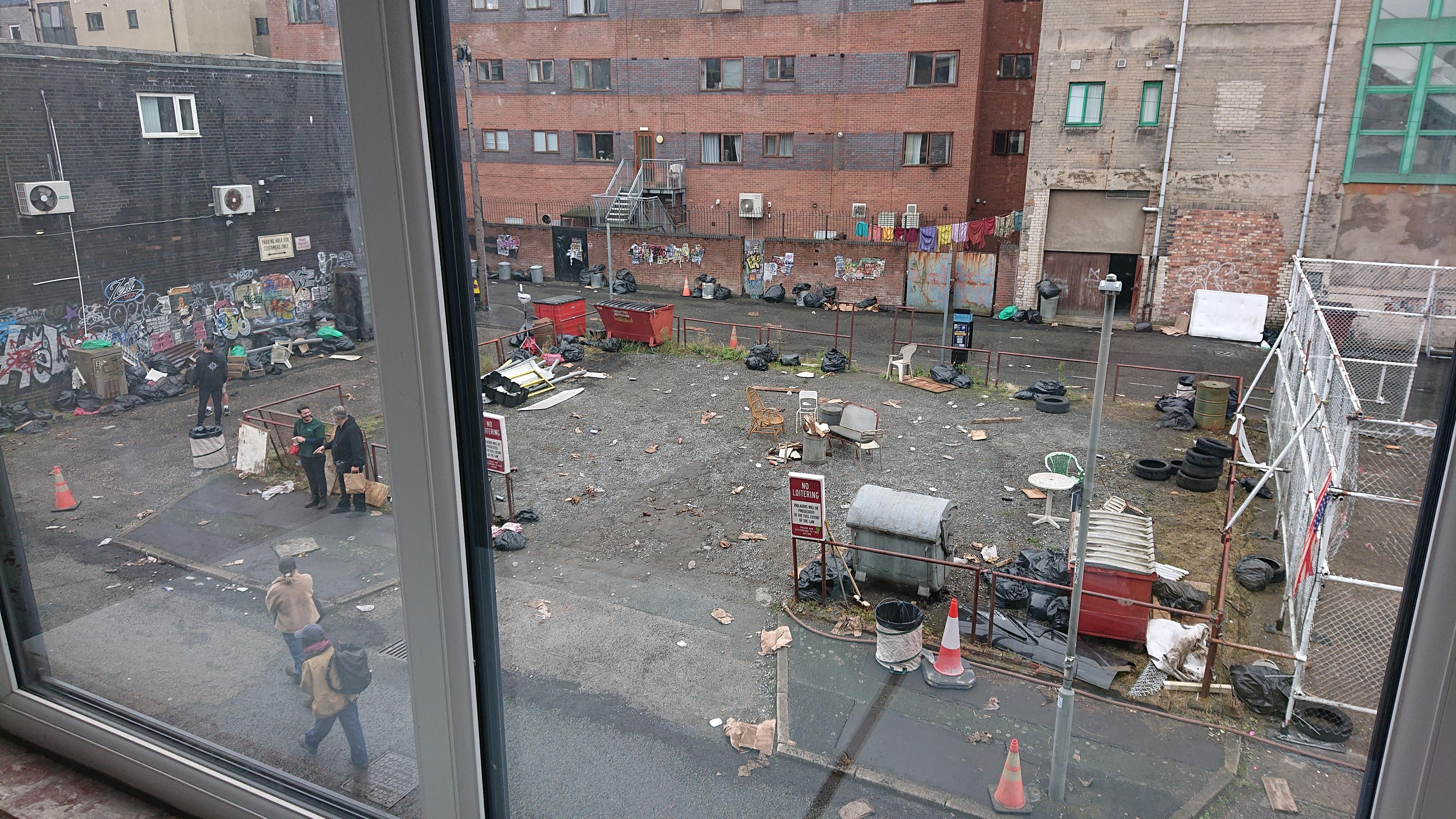 A gravel car park littered with carefully-placed bags of rubbish, some old chairs, piles of car tyres and other detritus. Dumpsters are car park's own (although then carefully weathered). There's some clothing hanging on a washing line outside a block of apartments that aren't actually being dried by the inhabitants. A few people can be seen milling around waiting for filming to start. If you look carefully, you can see one of Aeternum's air quality sensors mounted on the lamppost. We're not sure if it'll be in shot or not in the film (or if they'll edit it out in post)
