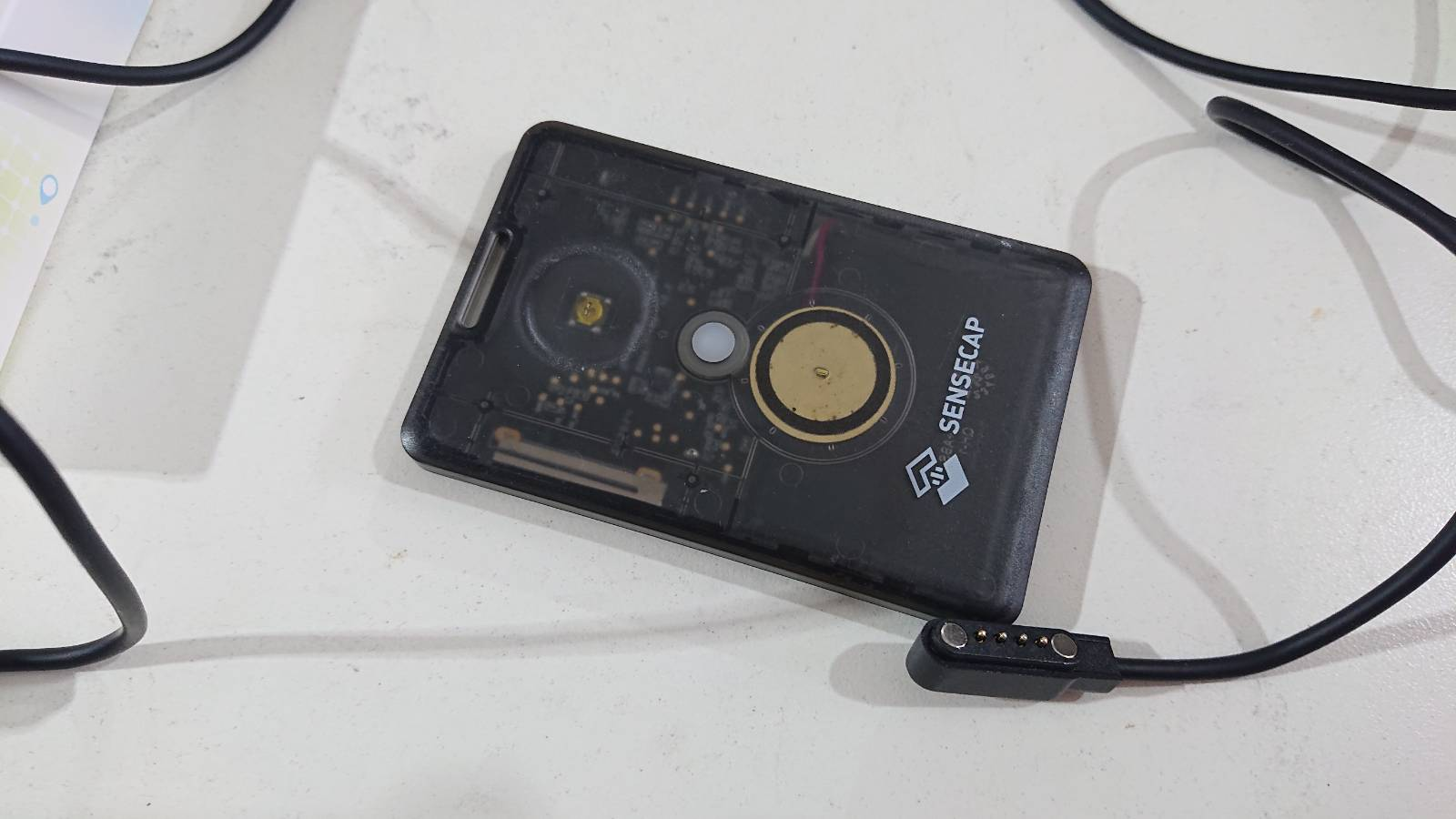 A Seeed Studio T1000-e Sensecap device, which looks rather like a thick credit card sits on a desk. The cable to charge/program it lies next to it. It's got a button and an LED on it (and a LoRa wireless modem, battery, GPS, etc. inside)