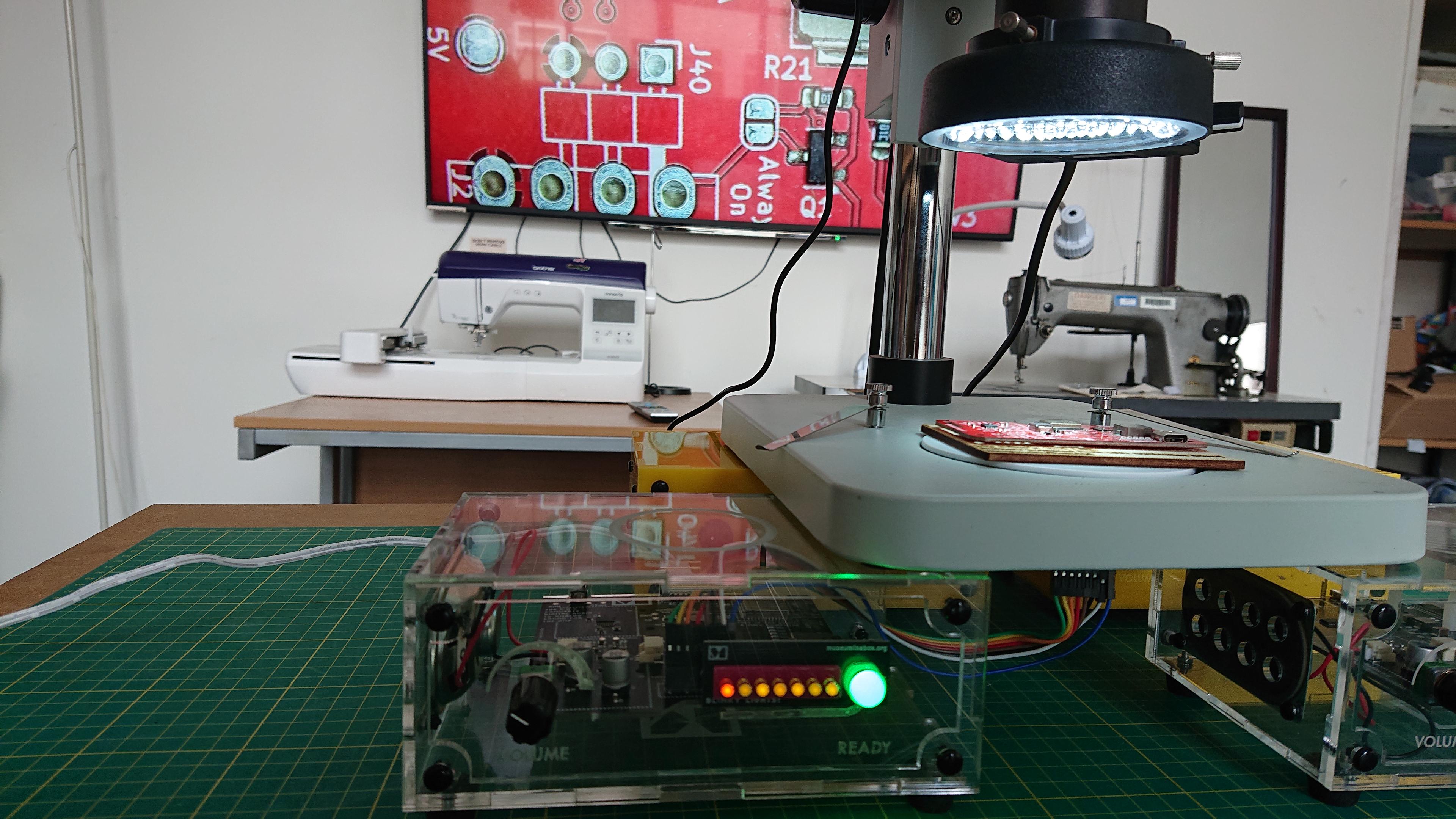 A microscope (we can only see the lower half of it) is sat upon a quartet of Museum in a Box boxes (look, we have them readily to hand). The Museum in a Box in centre of the frame is turned on (the LEDs on the front of it are all lit up) and some wires sneak out from the circuit board inside it to a position just under the stage of the microscope. There's a red circuit board (one of our My Bike's Got LED boards, as it happens) sat on the microscope stage. In the background there's a massive telly on the wall, and it's showing the output from the microscope: a close up of the red PCB. There's also a CNC embroidery machine and an industrial sewing machine in shot, but they're not important right now.