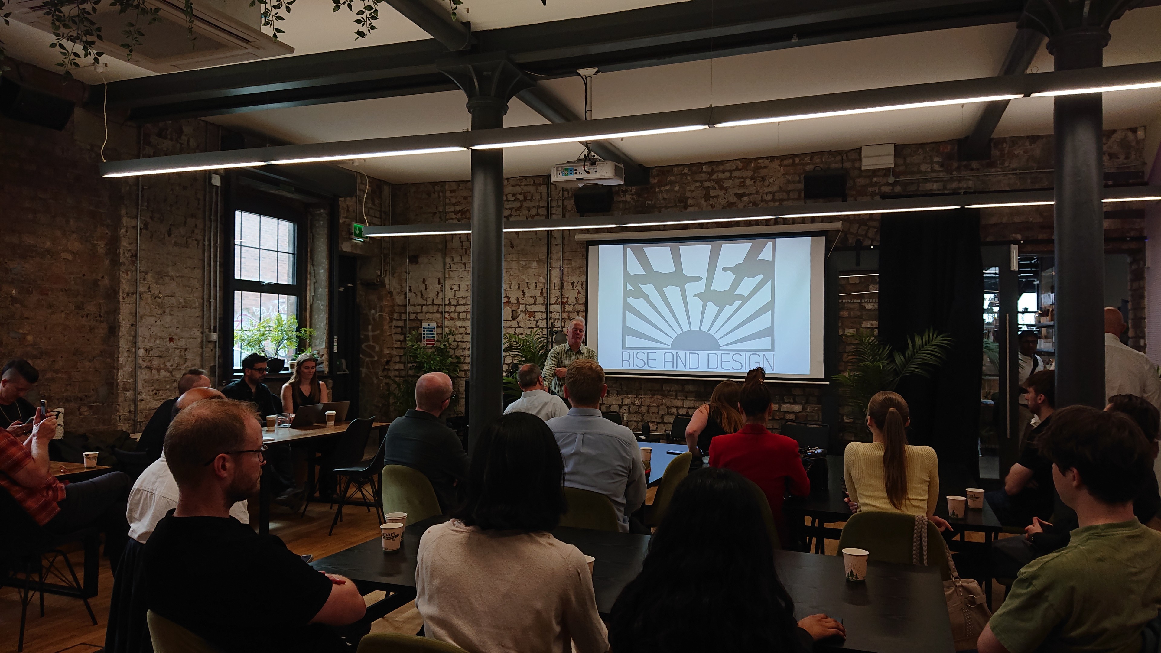 A room in an old converted brick warehouse where lots of people are sat at tables facing a projector screen which is showing a stylized sunrise above the words Rise and Design