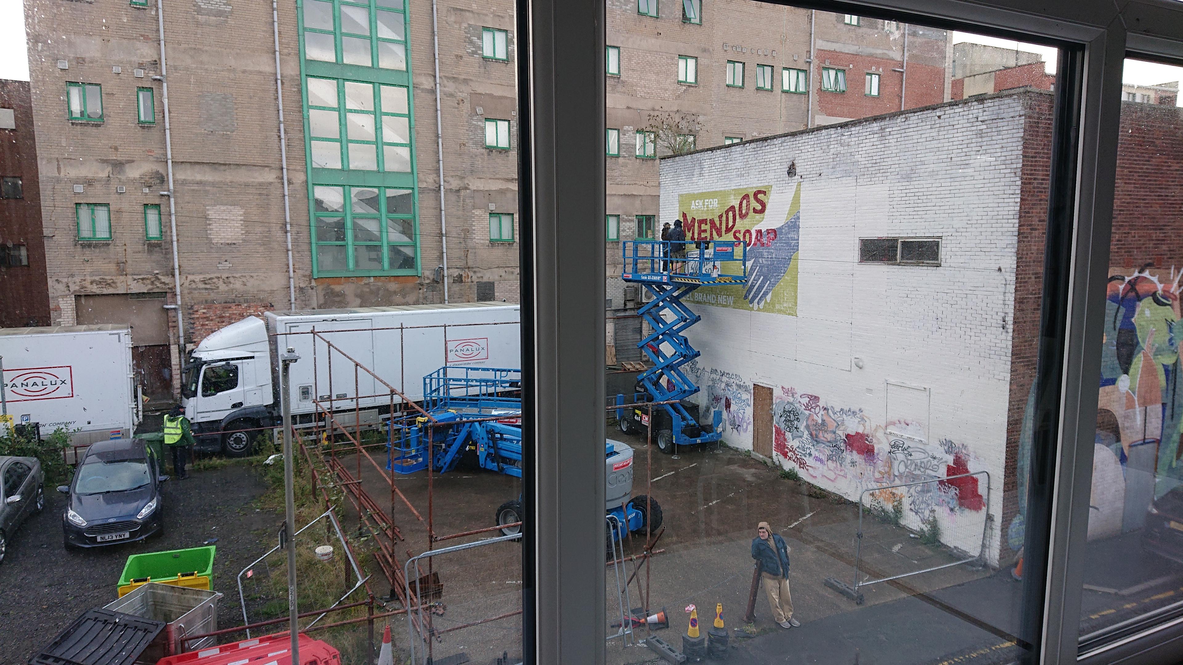 Looking out on an industrial back street in central Liverpool. There's a surface level car park next to a flat-roofed brick warehouse. The car park is devoid of cars and instead populated with a couple of blue cherry pickers and is surrounded by a high 