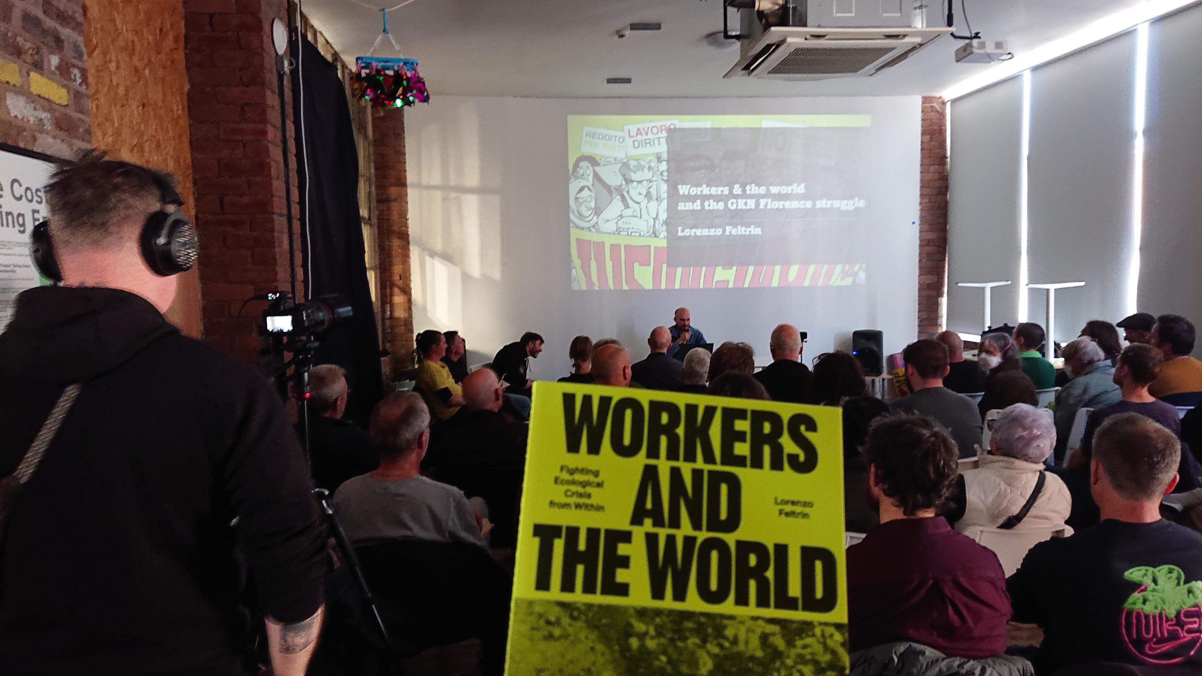 A copy of the book being held up in front of a view of a room full of people sat watching a presentation from Lorenzo