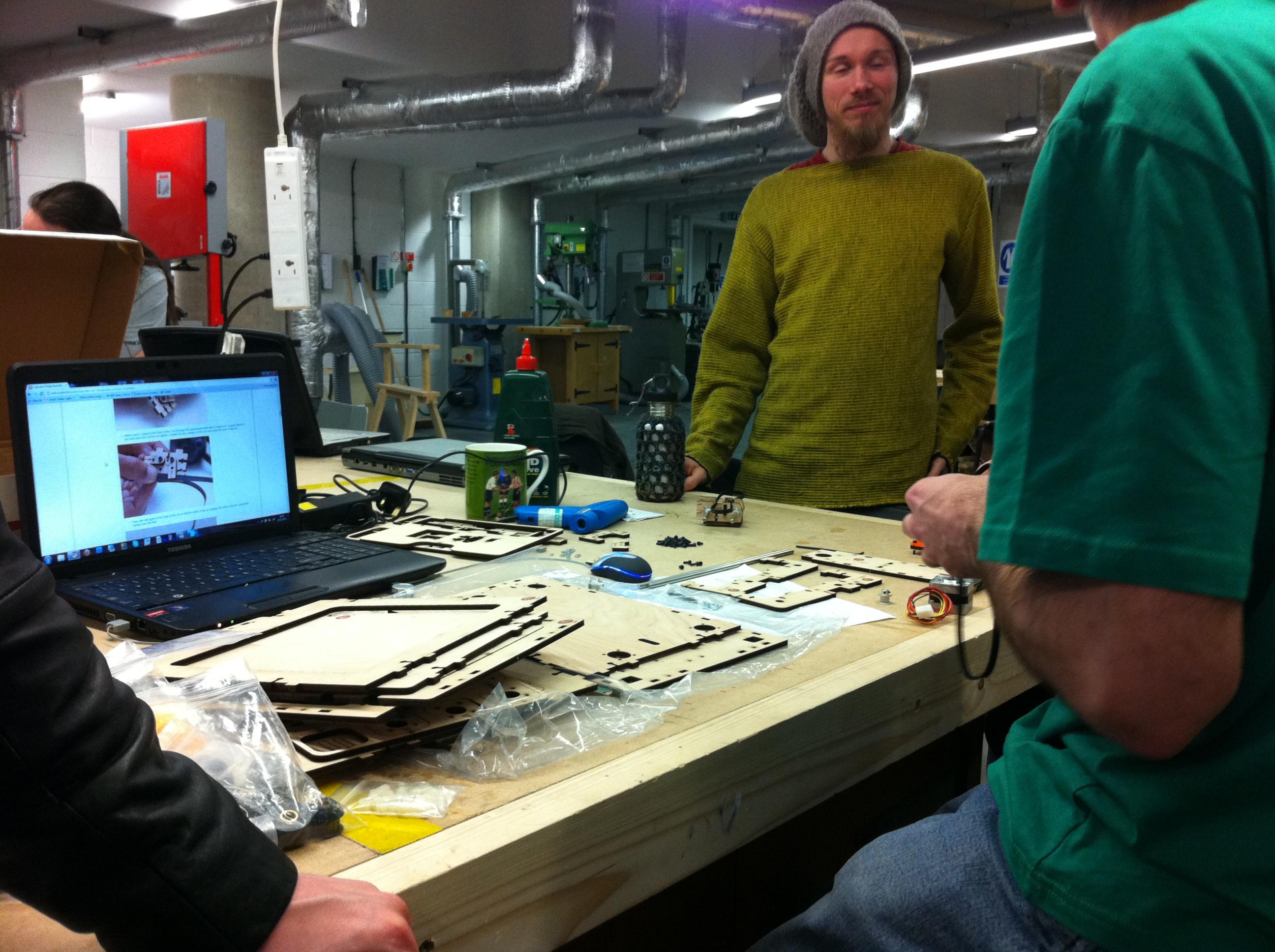 A couple of people are sat and stood around a workbench. The workbench has a stack of laser-cut wooden panels on it, and assorted bags of components. A stepper motor can just been seen over on one side. There's a laptop next to it all, showing the assembly instructions for a Makerbot Cupcake 3D printer