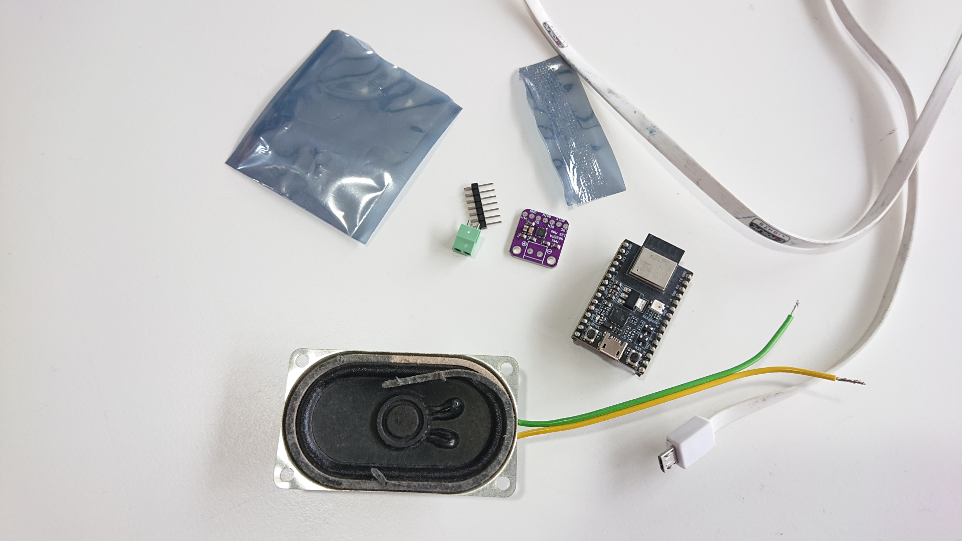 An array of components laid out on a desk. There's a small speaker, a little purple circuit board (that's an I2S DAC and class D amp) and an ESP32C3 breakout board. Plus a header and screw terminals to solder into the amp, the electrostatic bag it came in, and an USB cable