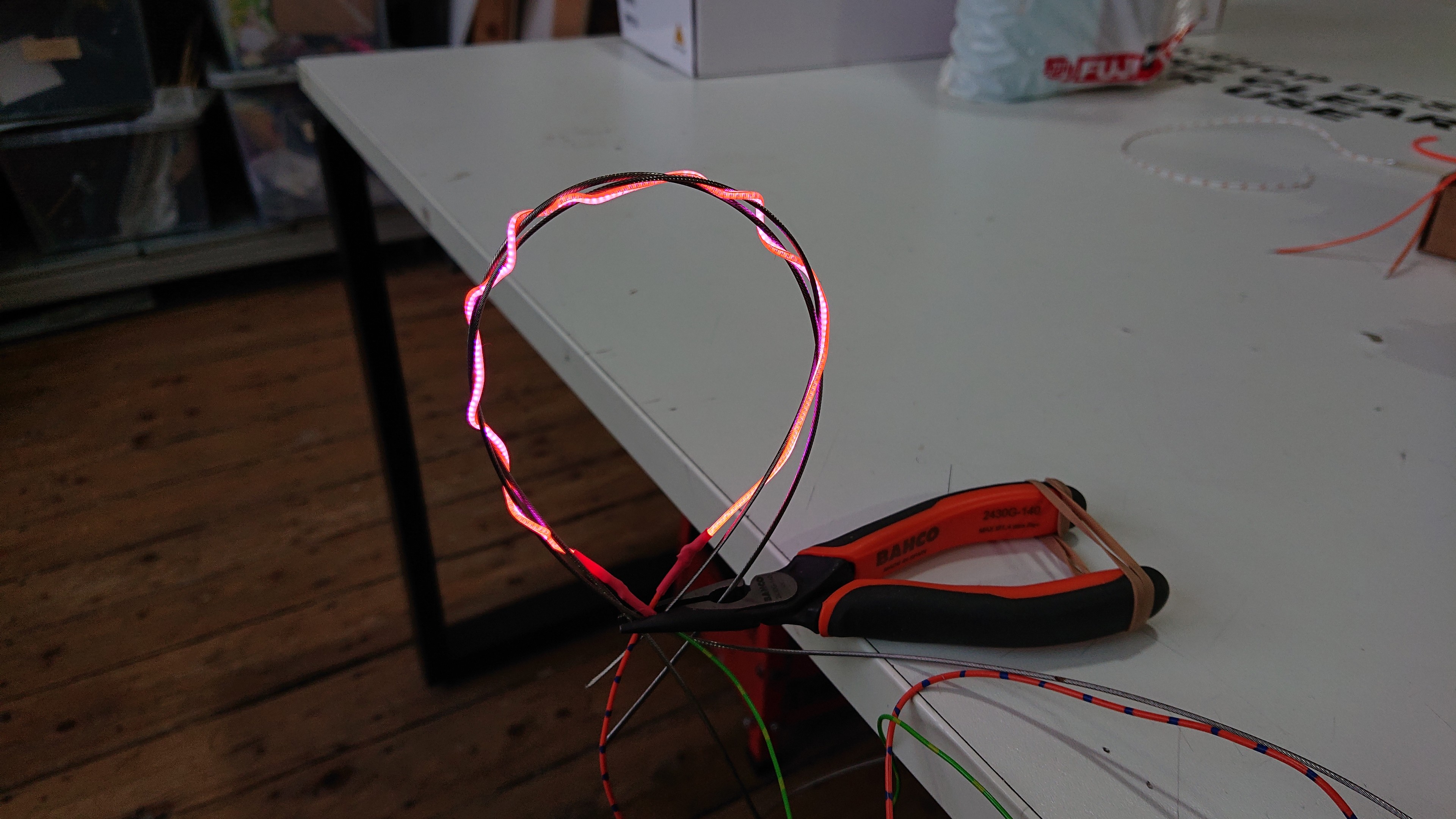 A loop made of plaited LED noodle and bike gear/brake cable, held by a set of long-nosed pliers. The handles of the pliers are resting on the edge of a table with the nose hanging over the edge. The loop is vertical in free air, and the LED noodle makes a wavy pattern due to the plaiting.