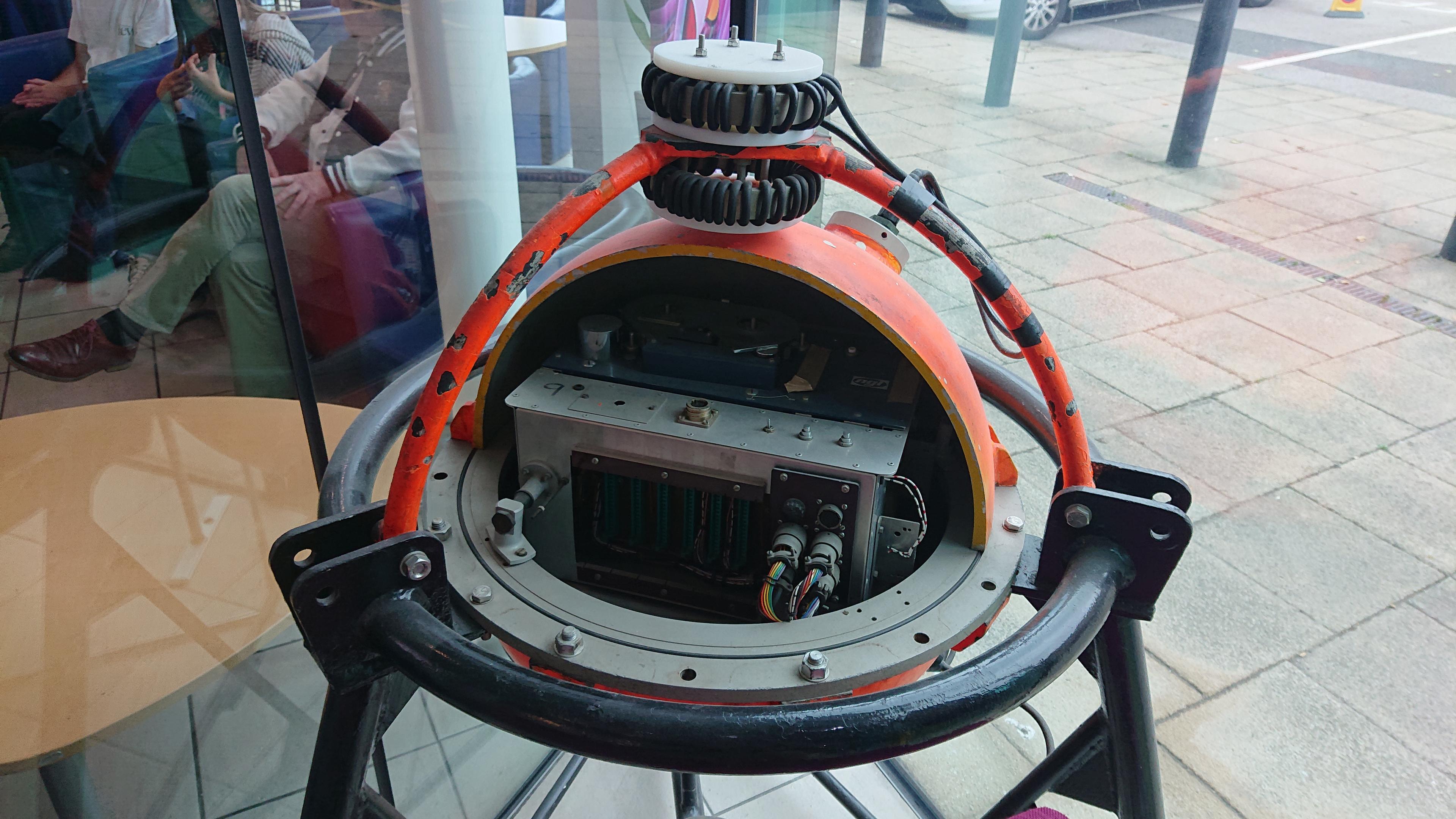 An orange-painted metal sphere, with a quarter of it cut away to show the internals - some cables and electronics. It is sat in a black tubular metal base, and has two toroid coils of cable (some sort on antenna maybe?) at the top