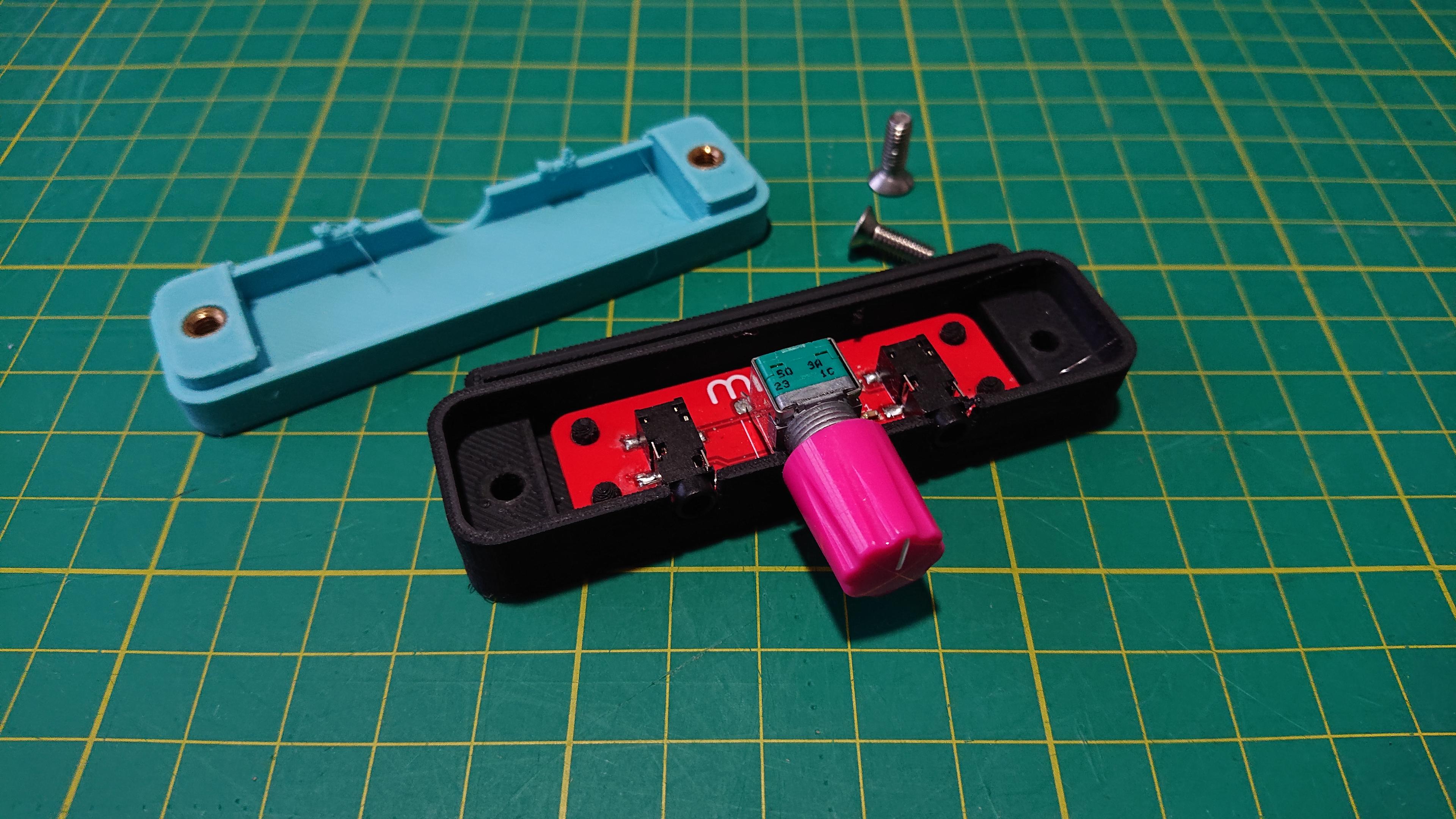 The case—split into its two halves—sits on a cutting mat. The upper half is teal, and shows the underside with the brass threaded inserts visible at either end. The lower half is black, and that has the red PCB with its stereo potentiometer and twin 3.5mm headphone sockets fitted inside on the mounting pegs. Next to both halves are a pair of countersunk M4 machine screws, ready to bolt it all together.