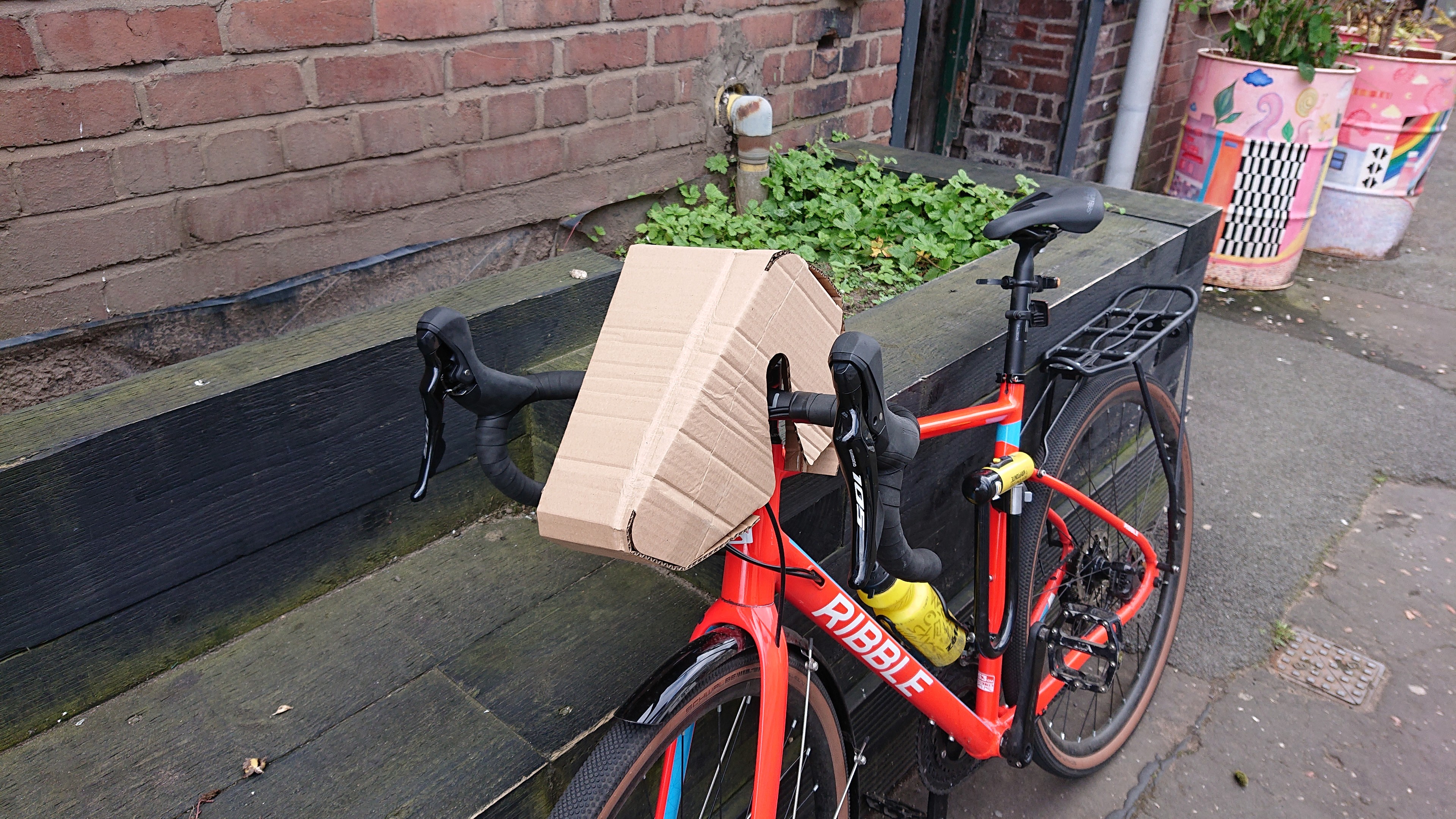 A gravel bike with drop handlebars leans against a planter outside a brick building. On the handlebars, looking a bit like a head for the bike, is the assembled brown cardboard wedge shape.