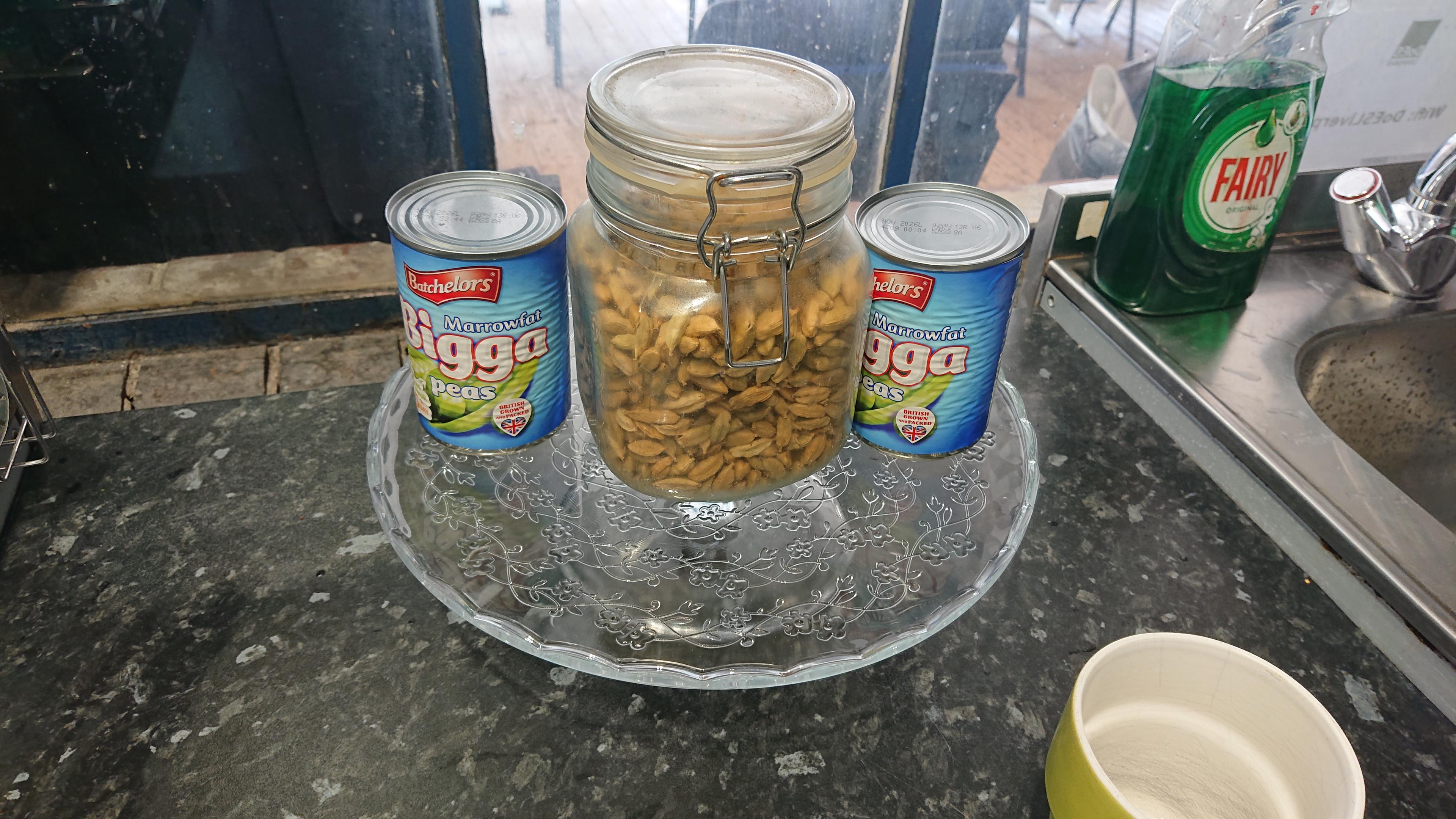 A fancy glass cake stand sits on the kitchen worktop in a shared office/makerspace. Rather than any cakes on it, there's a jar of some sort of nuts or seeds, flanked on either side by tins of marrowfat peas