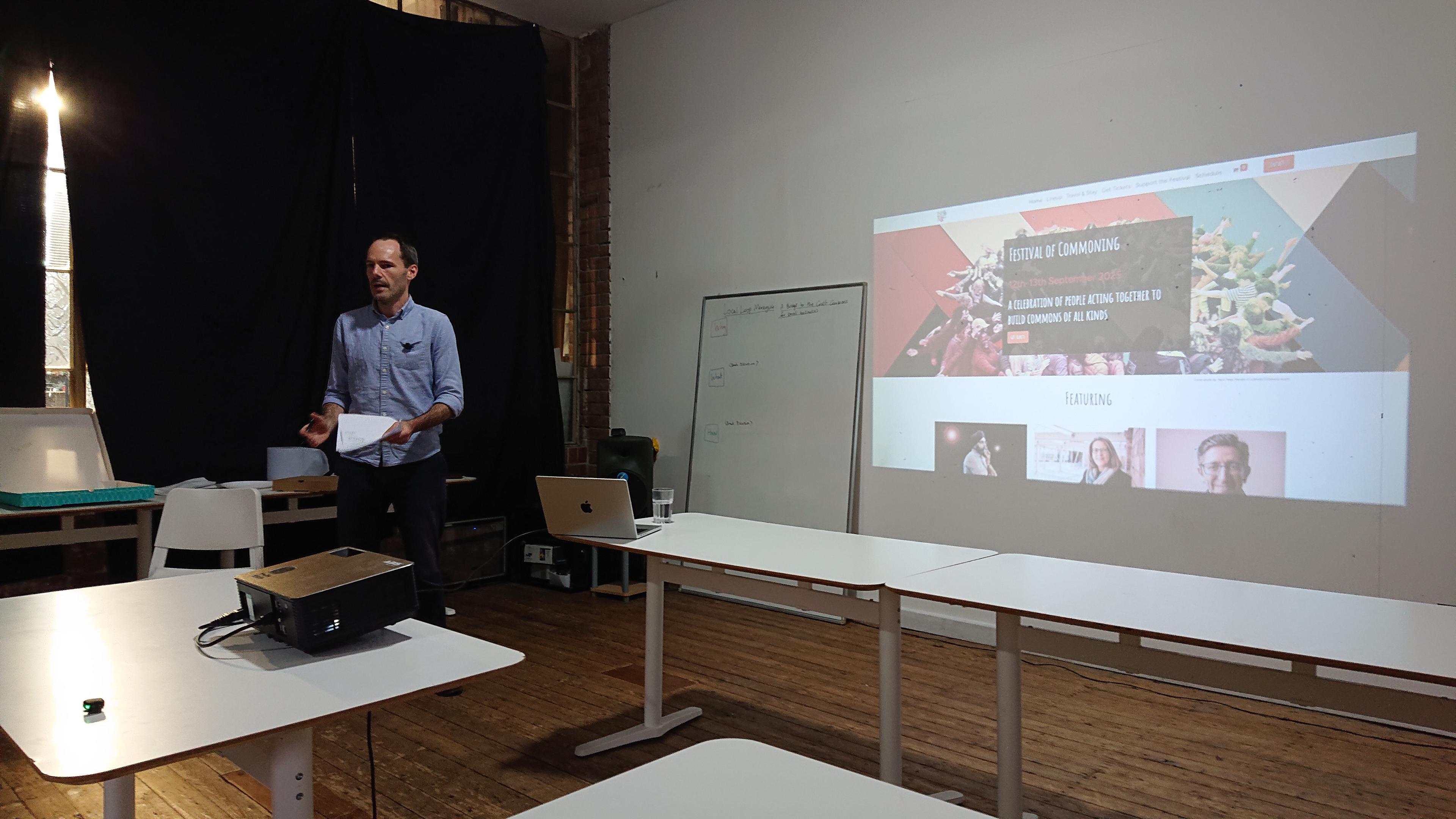 A man stands next to a projector screen at the front of the DoES Liverpool events room. The projector is showing the Festival of Commoning website