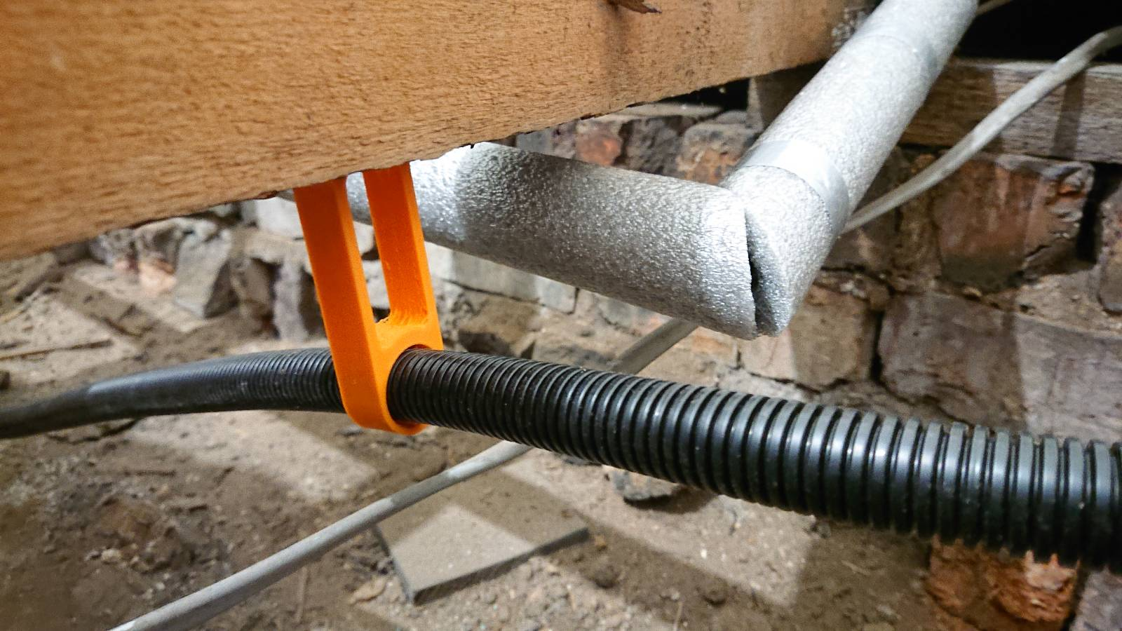 One of the clips installed under my dining room floor. It's screwed into the underside of the flooring joist and there's some black flexible conduit running through it. Running alongside is one of the pipes for the radiator, showing my not terrible but also not perfect mitred corner of the pipe insulation I've fitted. We can also see some of the low brick wall between rooms that the joist rests upon, and the bare dirt under the house, plus a grey mains cable.