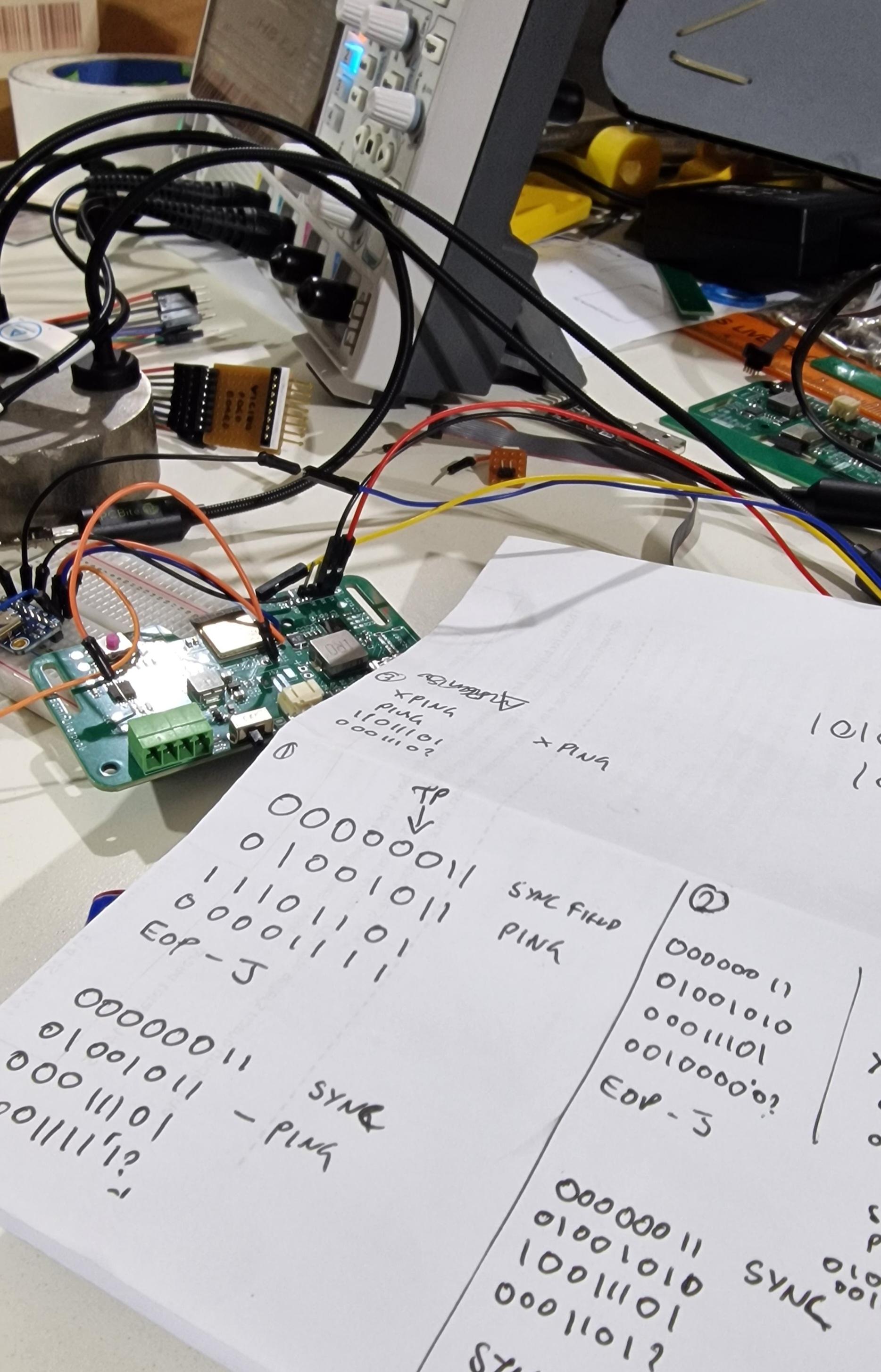 A very messy desk full of wires and circuit boards. Resting on top is a sheet of white paper scrawled with 0's and 1's. Highlights are '01001011 PING' AND '00000011 SYNC FIELD'