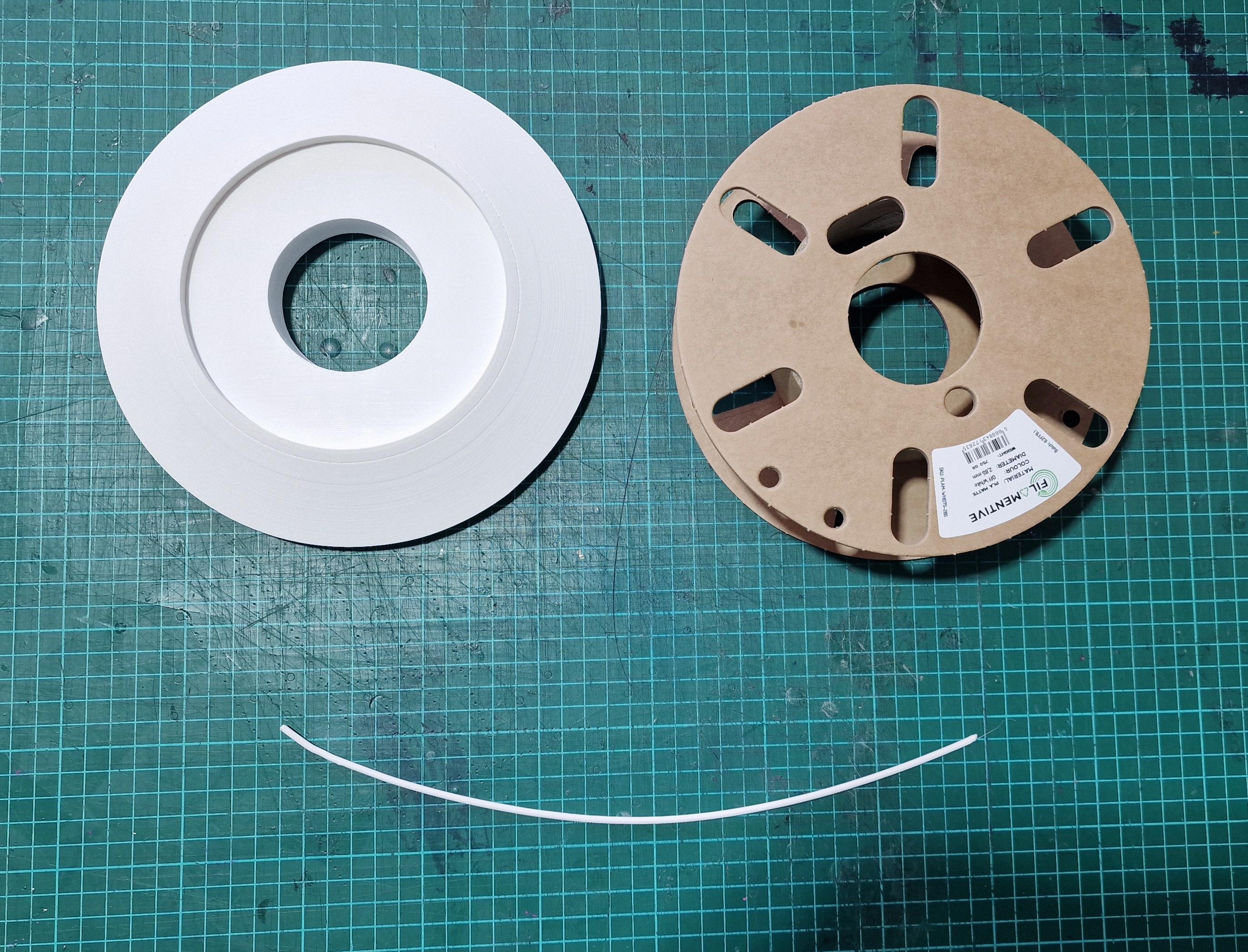 Green cutting mat with a white circular 3d model, and empty spool and a short curve of filament together making a smiley face.