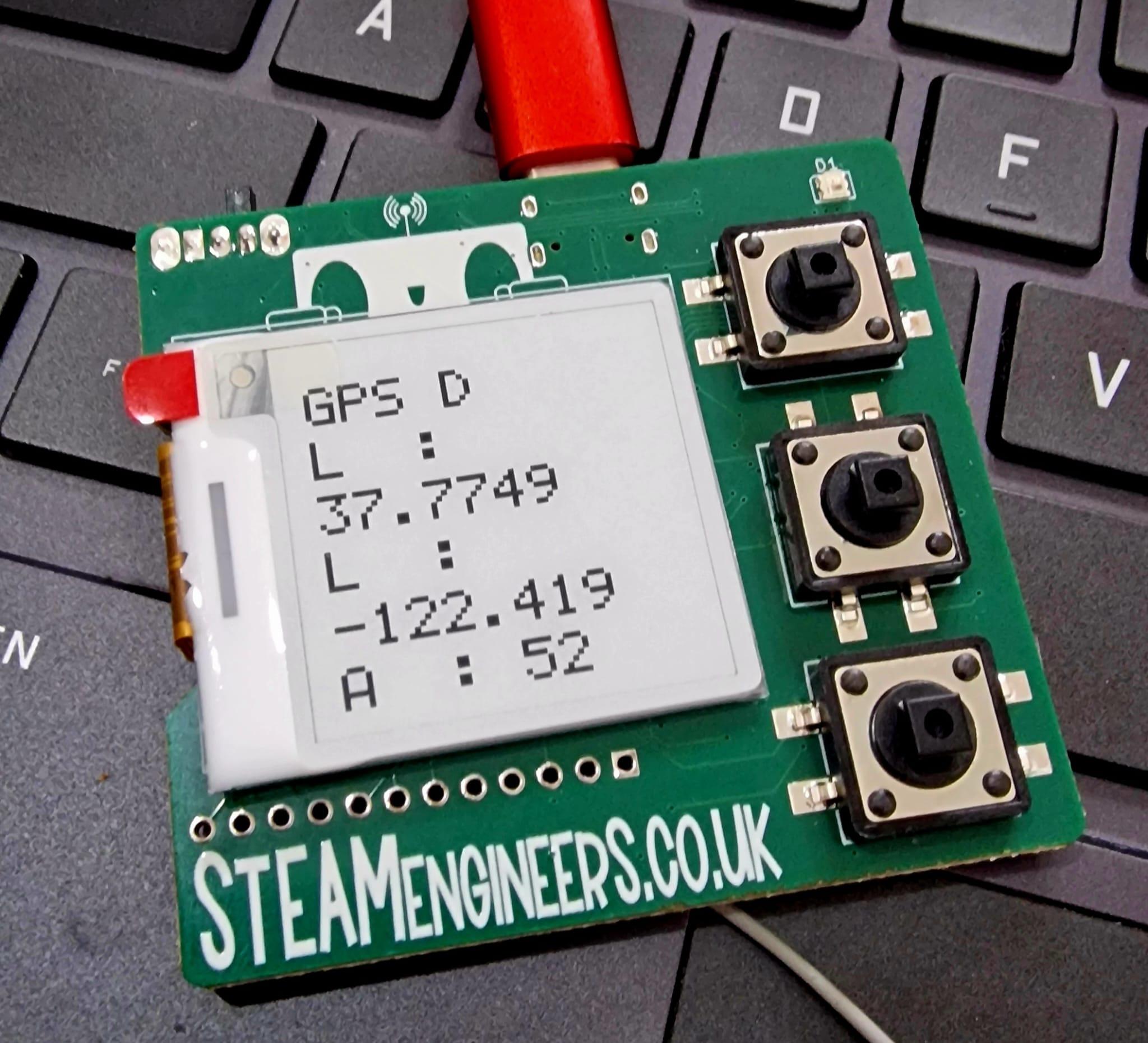 A green pcb sat on a laptop keyboard. On the board is a 25mm square eink screen showing dummy gps data. Above the screen on the silkscreen is a picture of a robot peering over. To the right are three black buttons. Below the screen is the legend STEAMEngineers.co.uk 