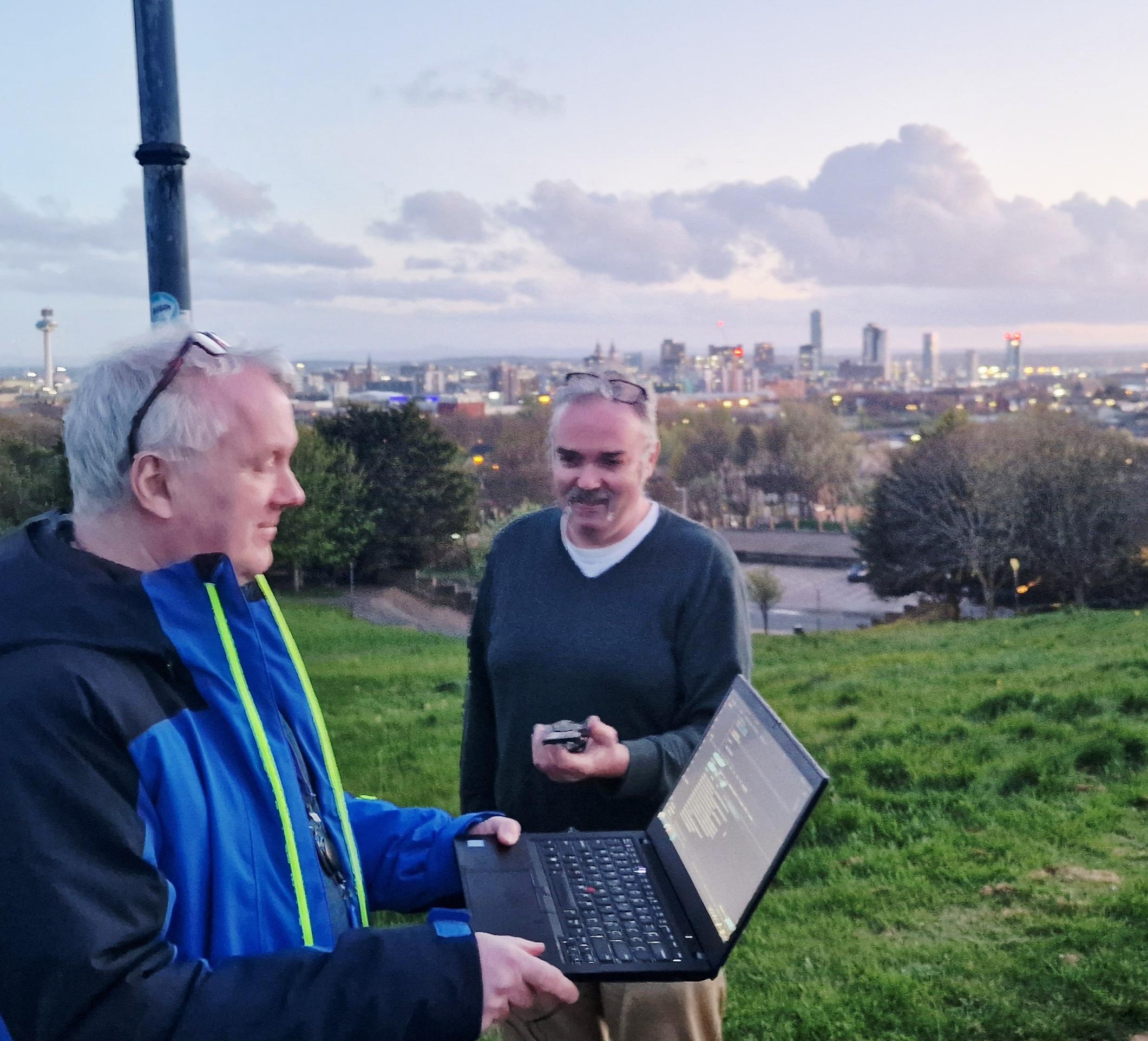 In thr foreground two people are stood on the top of Everton brow, behind them is the Liverpool skyline. One is holding a laptop reading serial output from the pcb the other is holding. It managed to find 30+ satellites to listen to and determined where it was very well. Everyone, including the unseen camera man is smiling at the result.