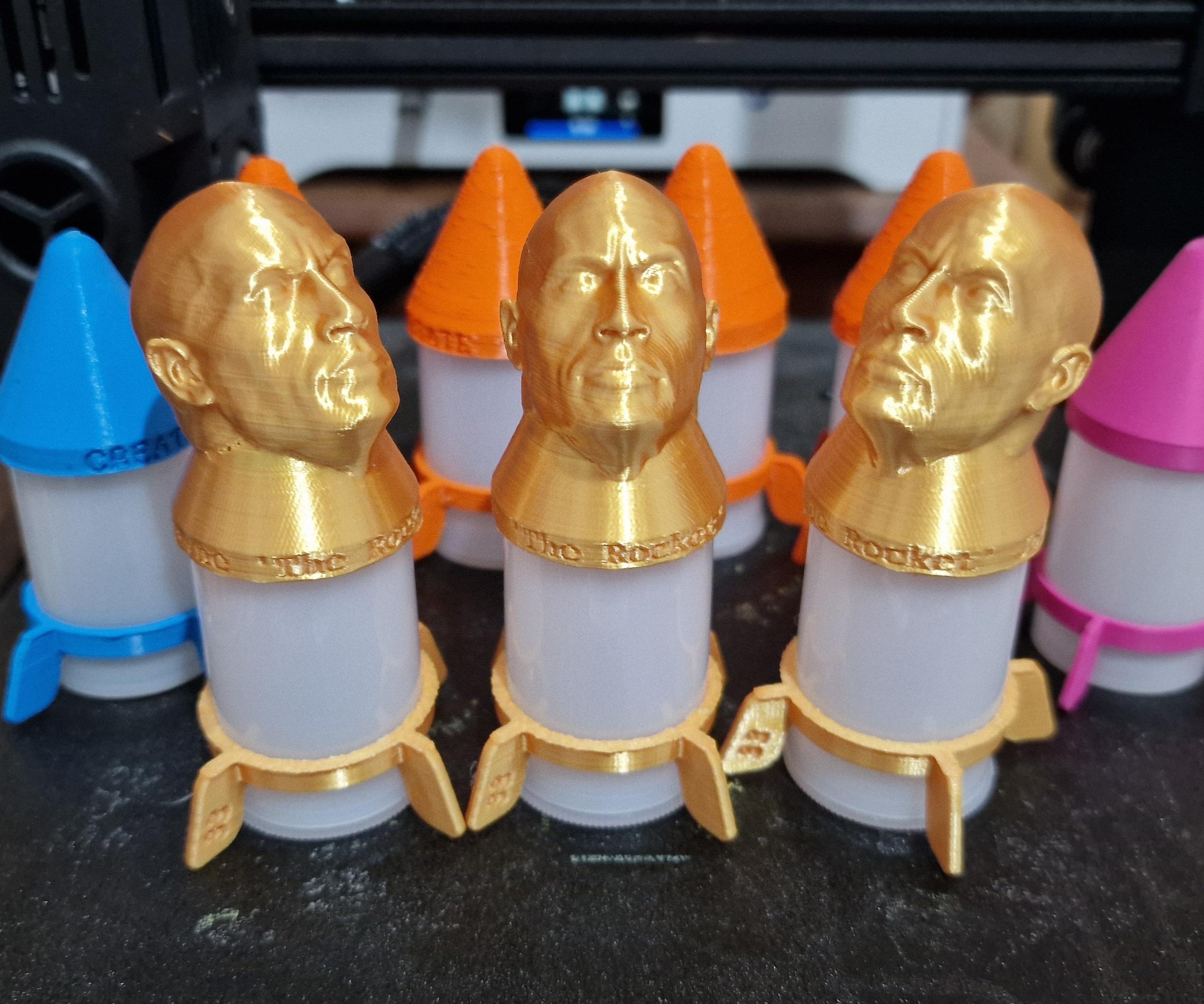 A collection of 3d printed 35mm film canister rockets. In the background standard rocket shaped rockets in orange, pink and blue. In the foreground three golden rockets, each with Dwayne the rock Johnsons head on top of the nose cone. Sometimes the why questions aren't that important.