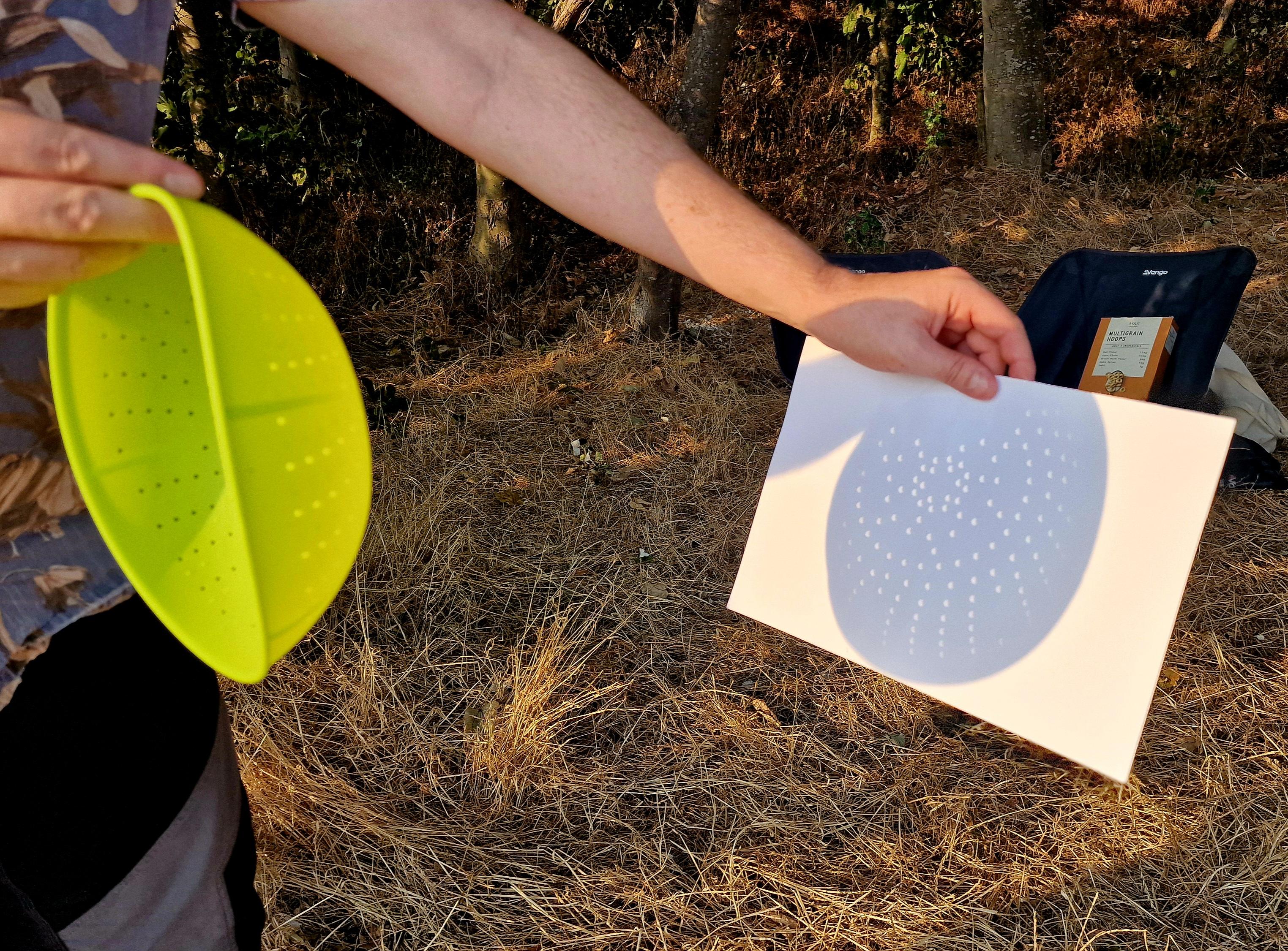 A man holding a yellow colander, projecting multiple images of a solar eclipse onto a sheet of white paper.