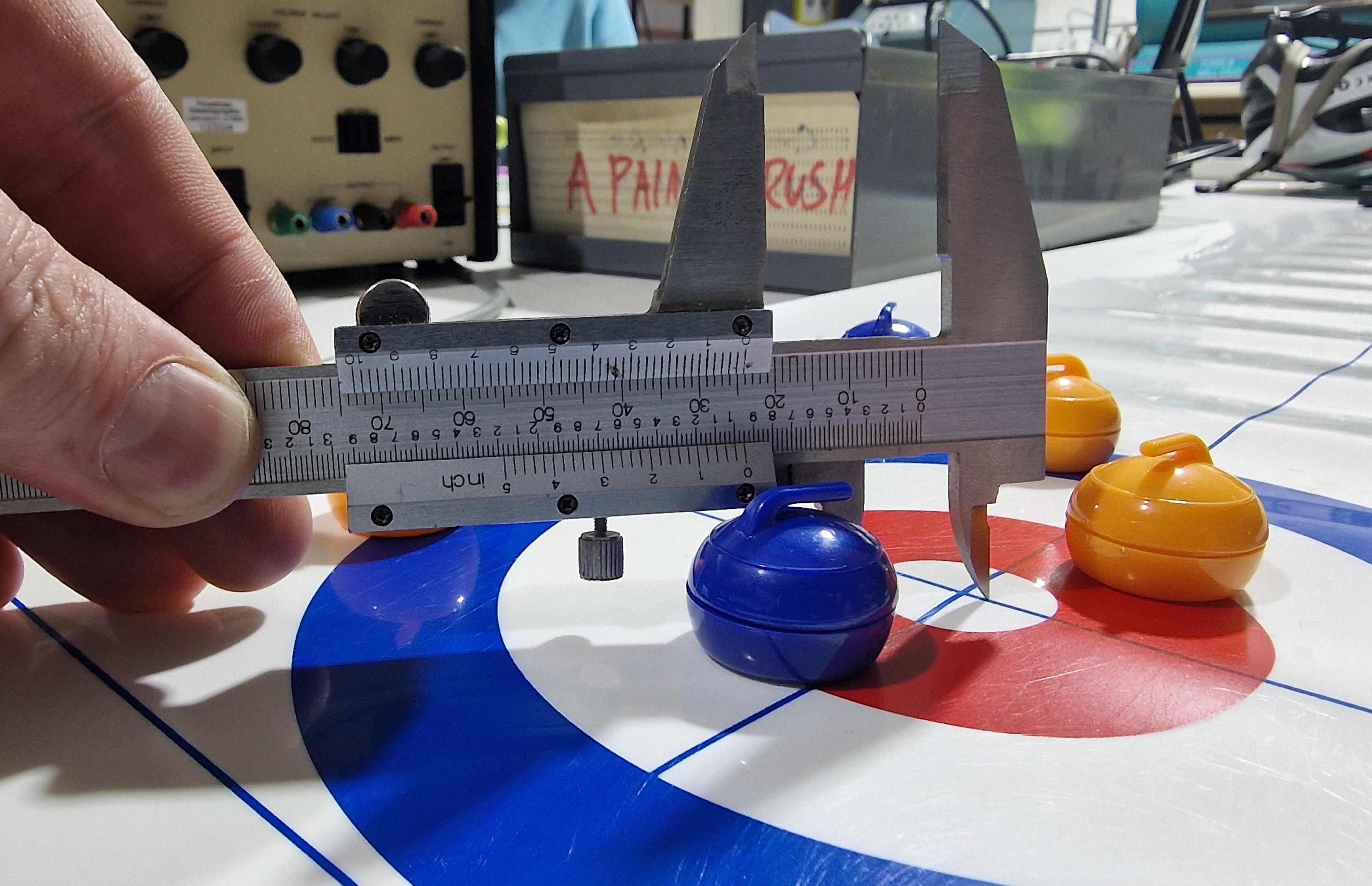 Image shows a table top curling game with mini blue and yellow curling stones in a blue, white and red house. Vernier calipers are being used to measure which is closest.