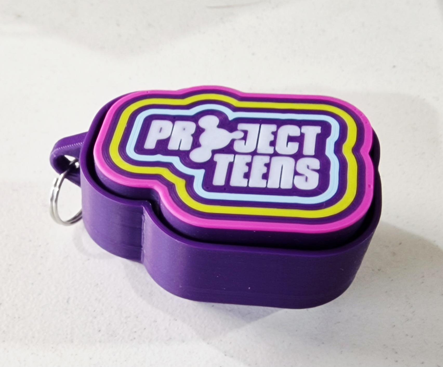 A five coloured 3d print of the design. The bottom piece is in purple and has a key chain for the important rucksack attachment. The top, clicker piece is printed in purple, the project teens logo is in white and it's surrounded by light blue, yellow and pink bands. 